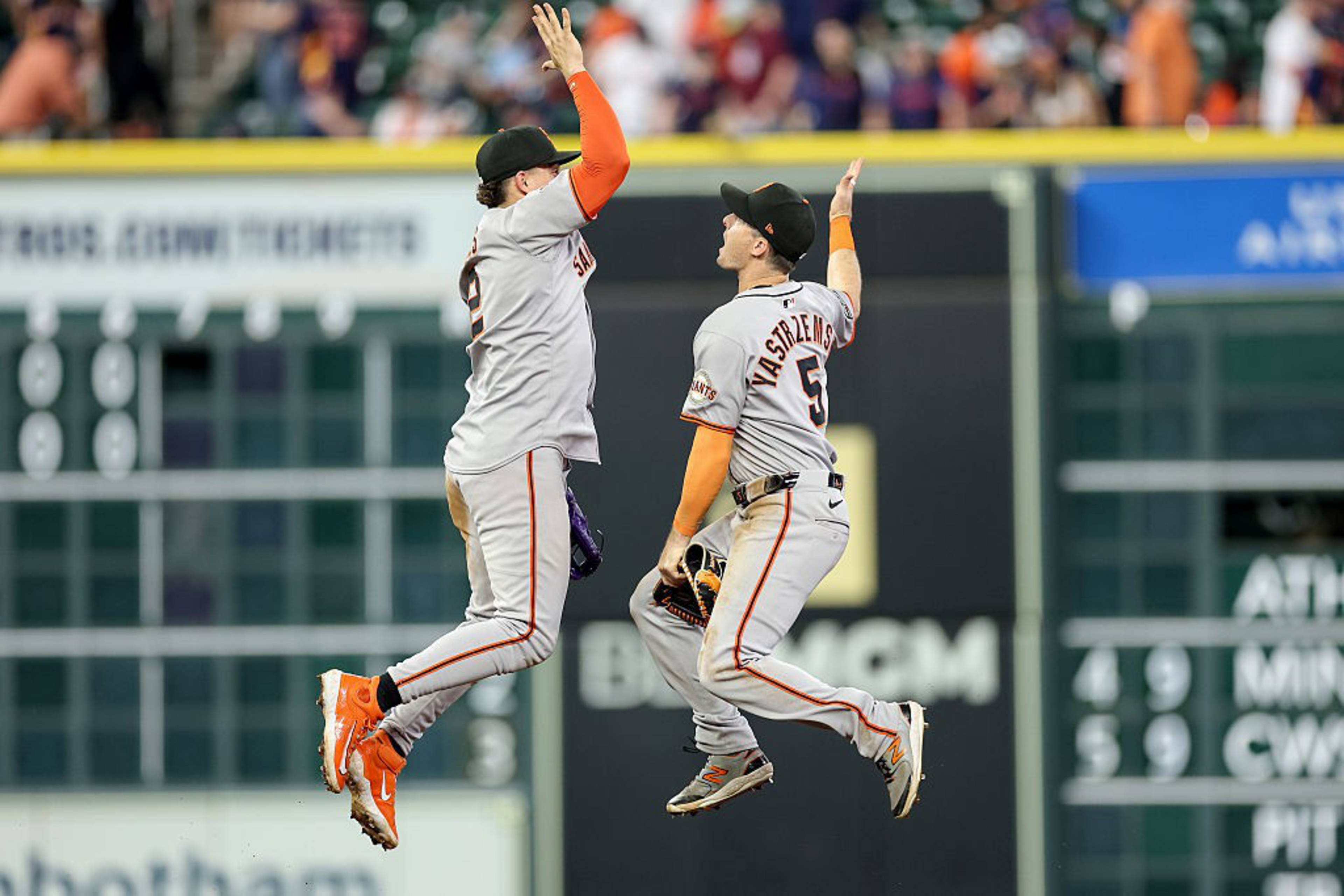 Two baseball players in gray uniforms perform a celebratory jump and high-five on the field, with a blurred audience in the background.