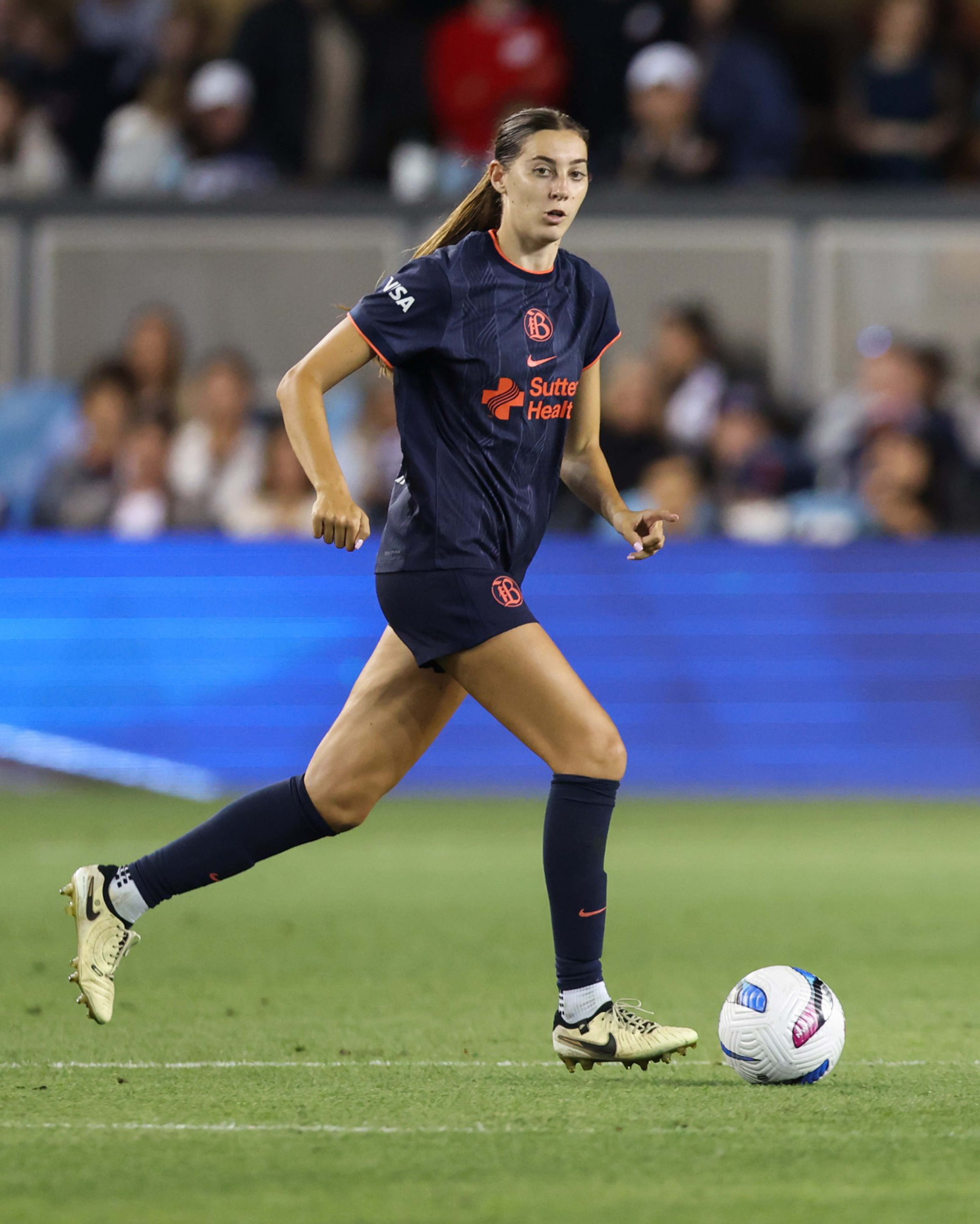 A female soccer player in a dark blue uniform with orange accents runs on the field, focused on a white soccer ball near her feet.