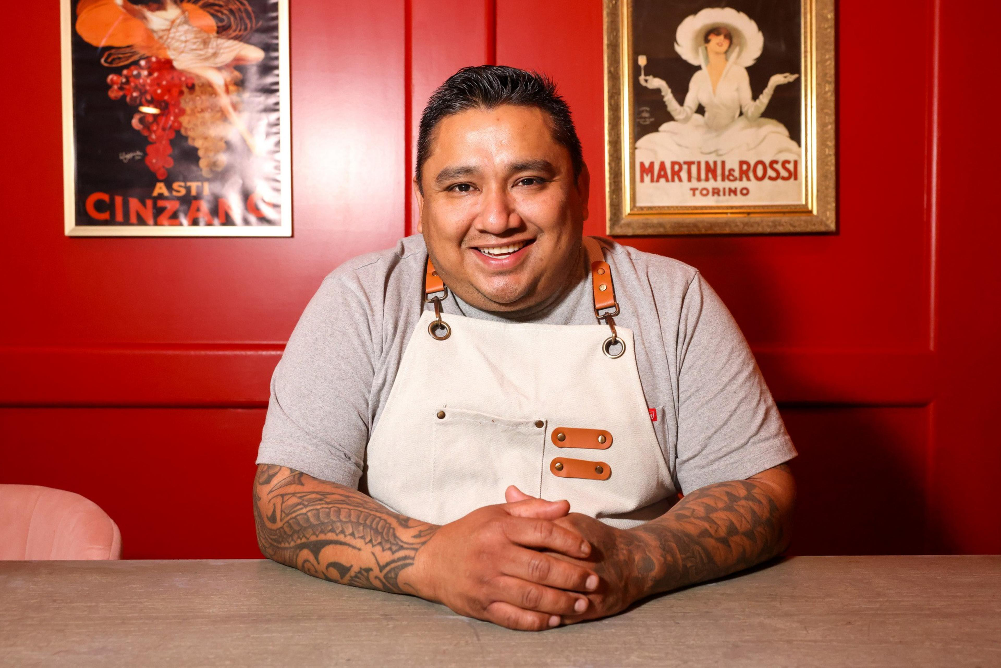 A smiling person with tattooed arms sits at a table, wearing a gray shirt and a white apron against a red background with vintage posters.
