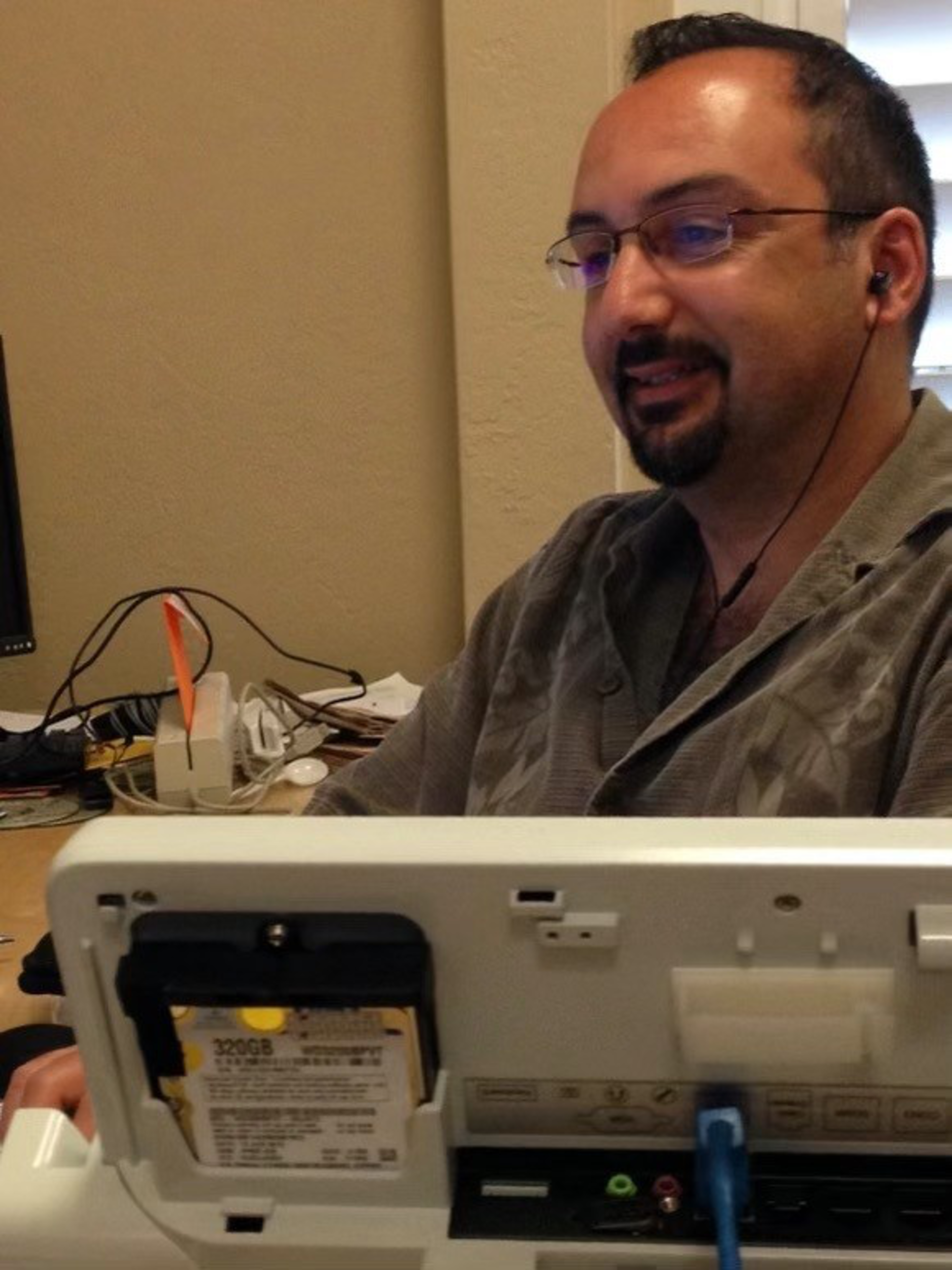 A man with glasses and earbuds is smiling while sitting at a desk, facing a computer with its back panel open showing a 320GB hard drive.