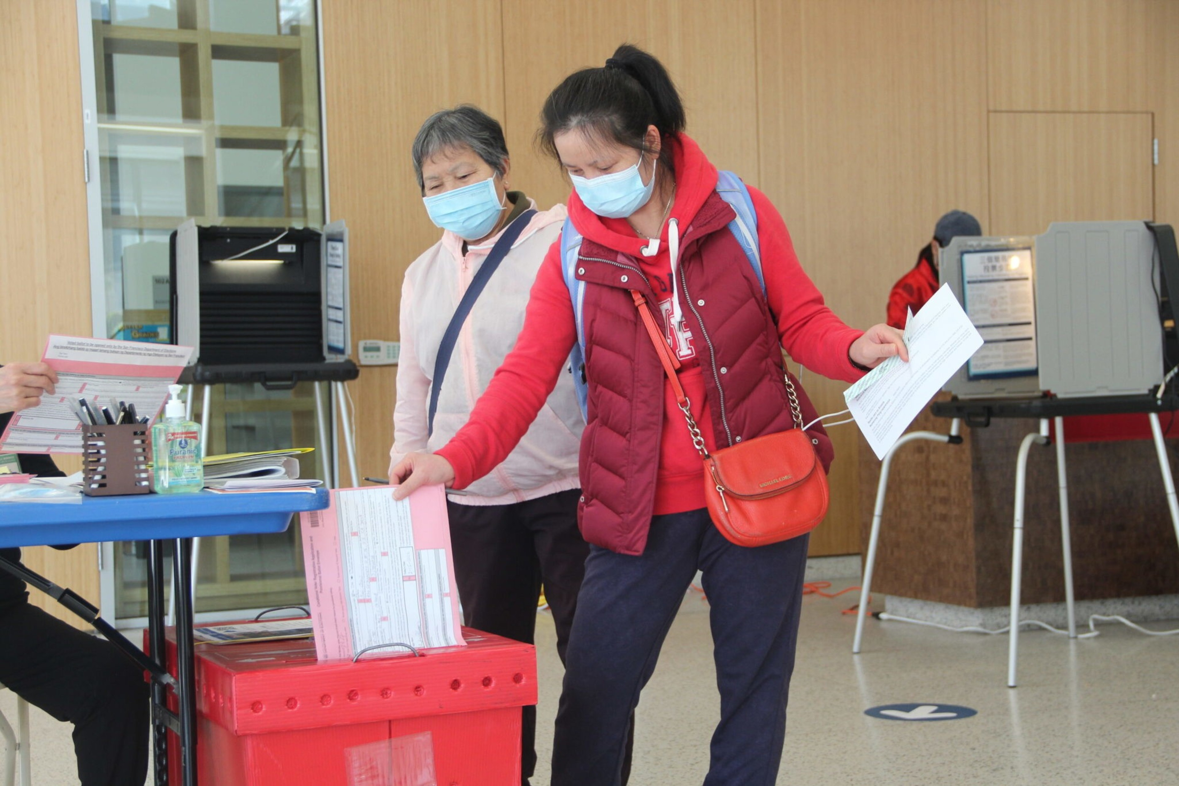 Two masked women stand near a table and a red ballot box, with one woman casting a paper vote while holding documents.