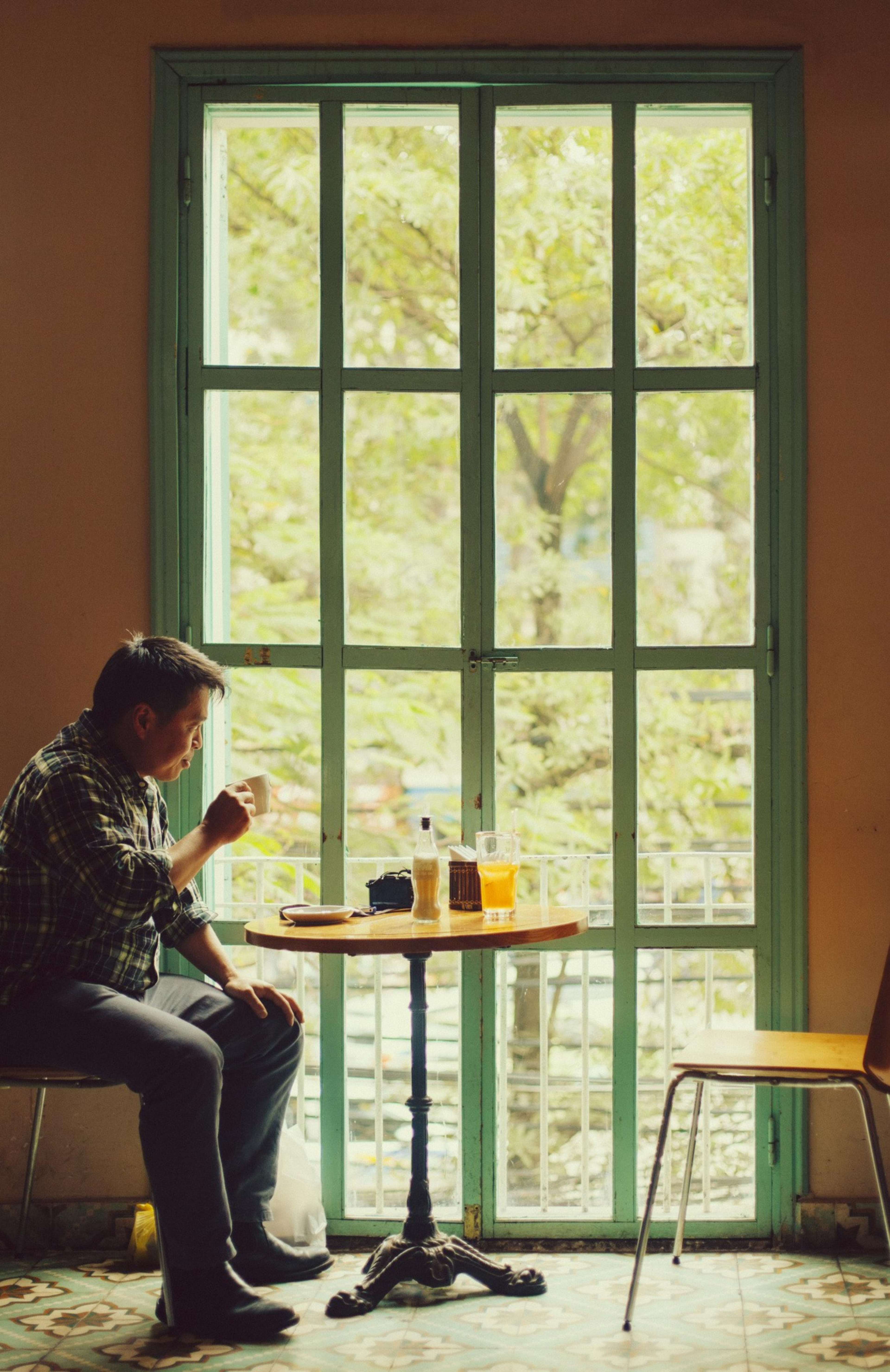A person sits at a small wooden table by a large window, sipping a drink. The table holds a bottle and a glass of orange juice, with a leafy view outside.