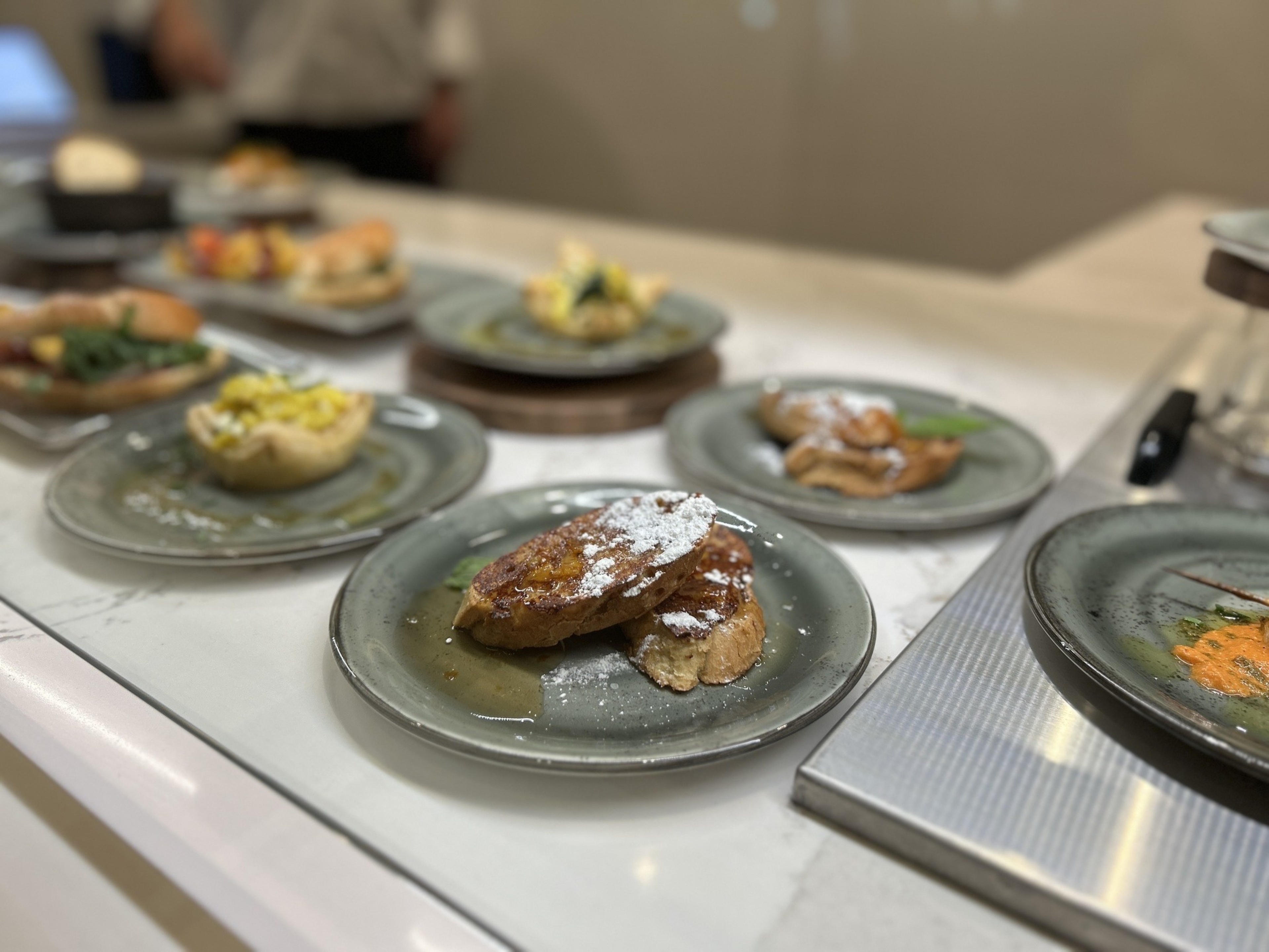 The image shows several plates of various dishes on a table. The foreground has French toast with powdered sugar. Other plates feature diverse, colorful foods.