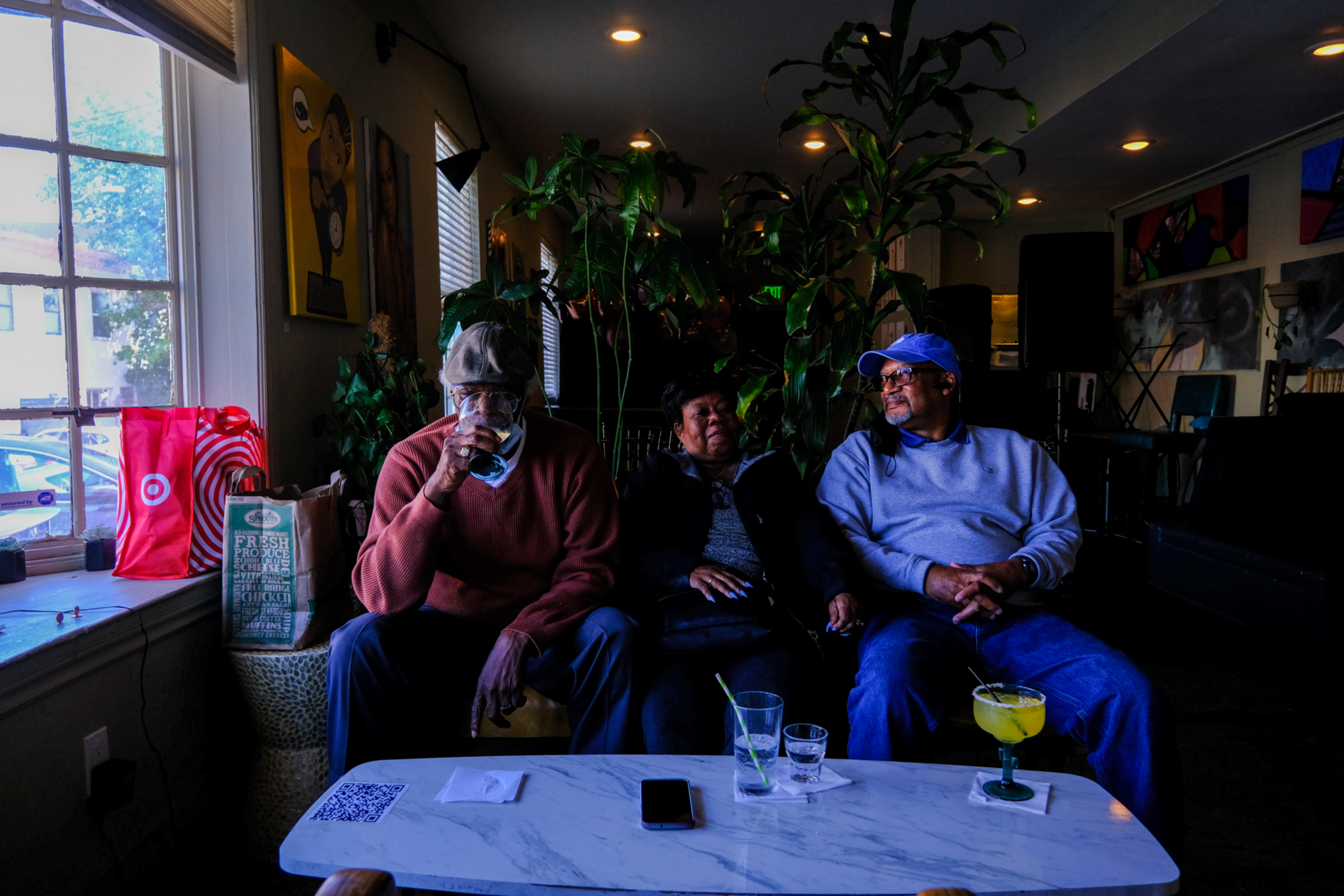 Three people sit on a couch in a dimly lit room, with drinks on a table in front; green plants and colorful paintings decorate the background.
