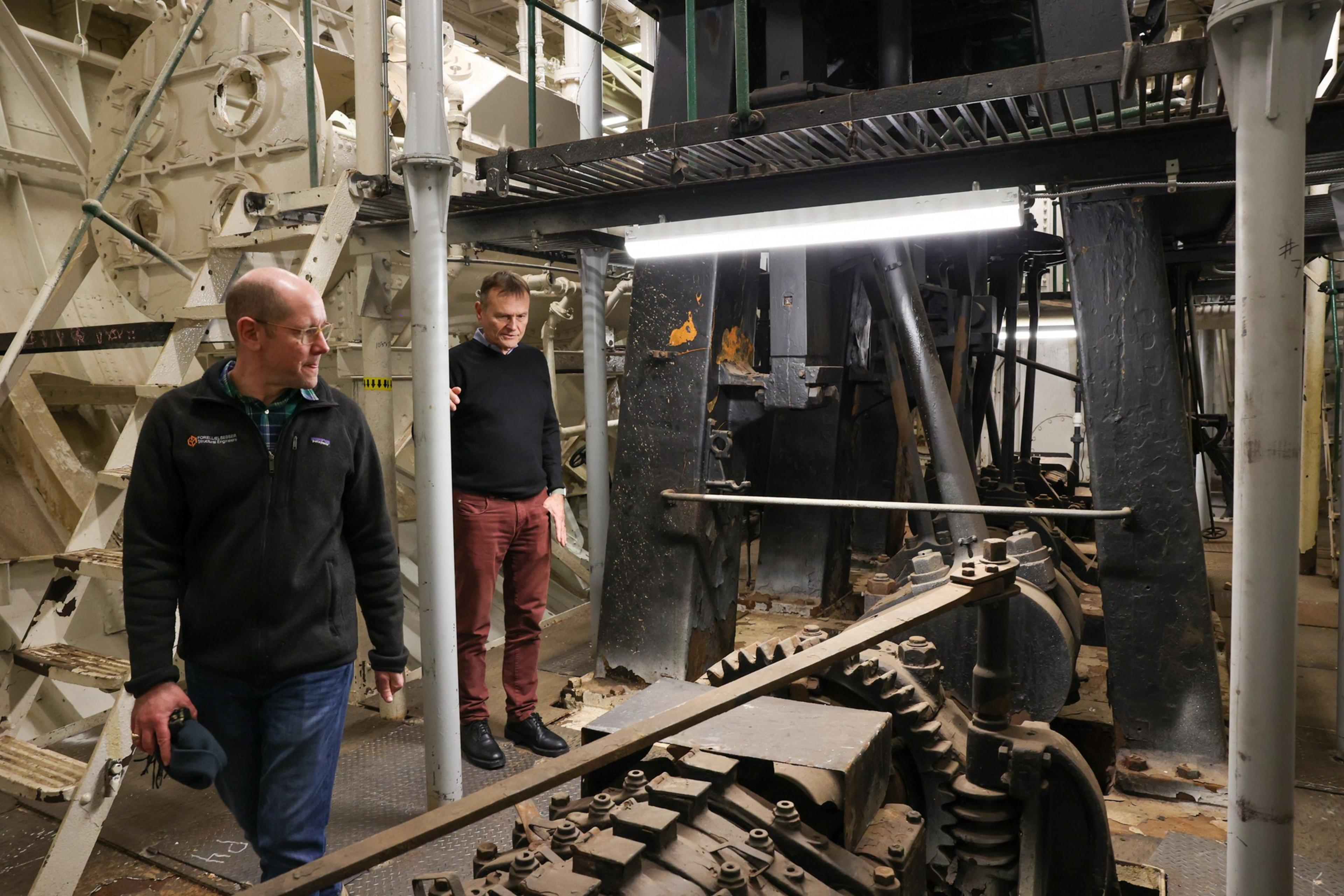 Two men stand amidst large, old industrial machinery with rusted metal gears and beams inside a weathered, lighted factory or workshop space.