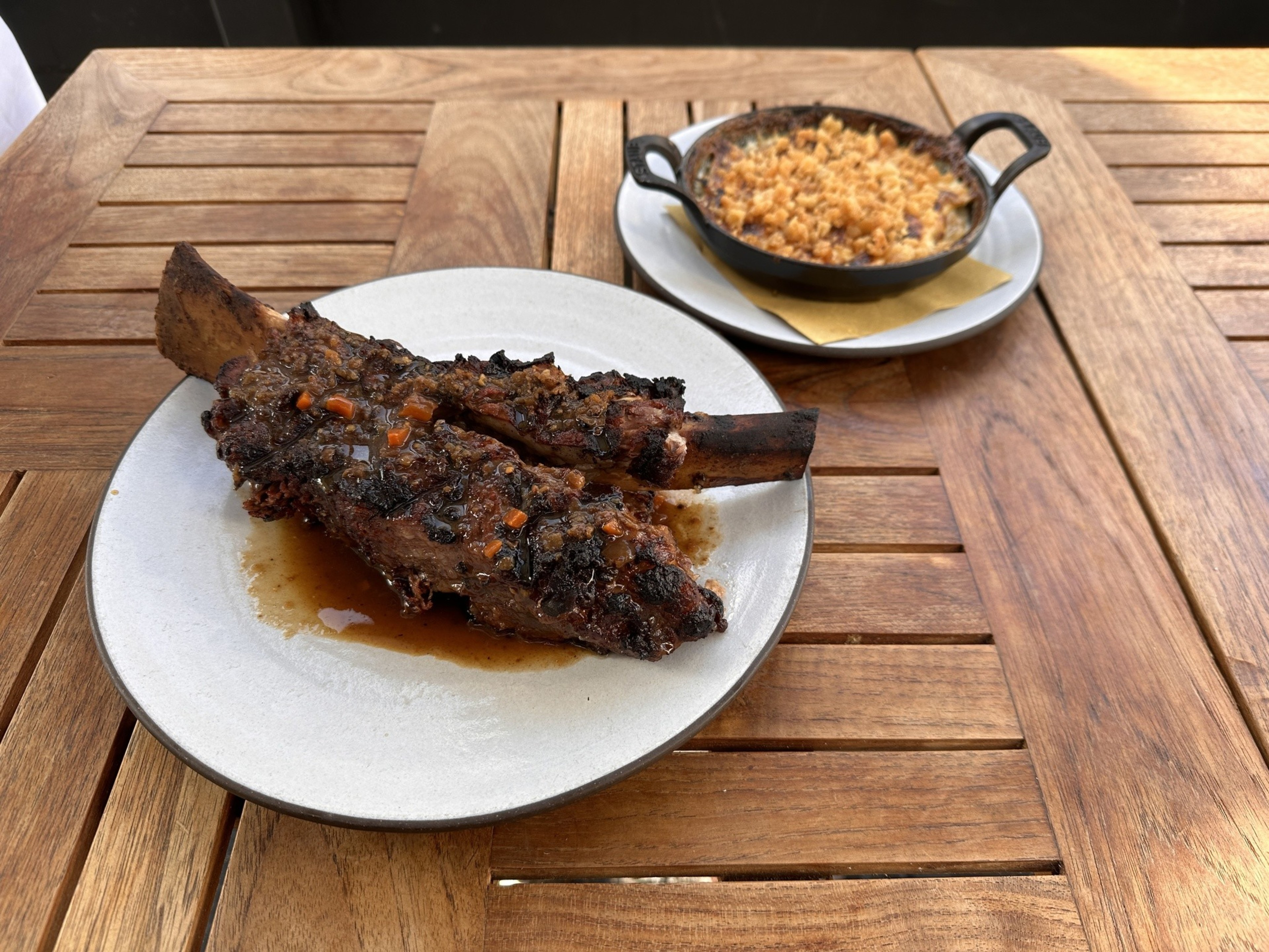 A wooden table holds a plate of juicy, glazed ribs with vegetables, next to a small cast iron pot filled with a macaroni and cheese dish.