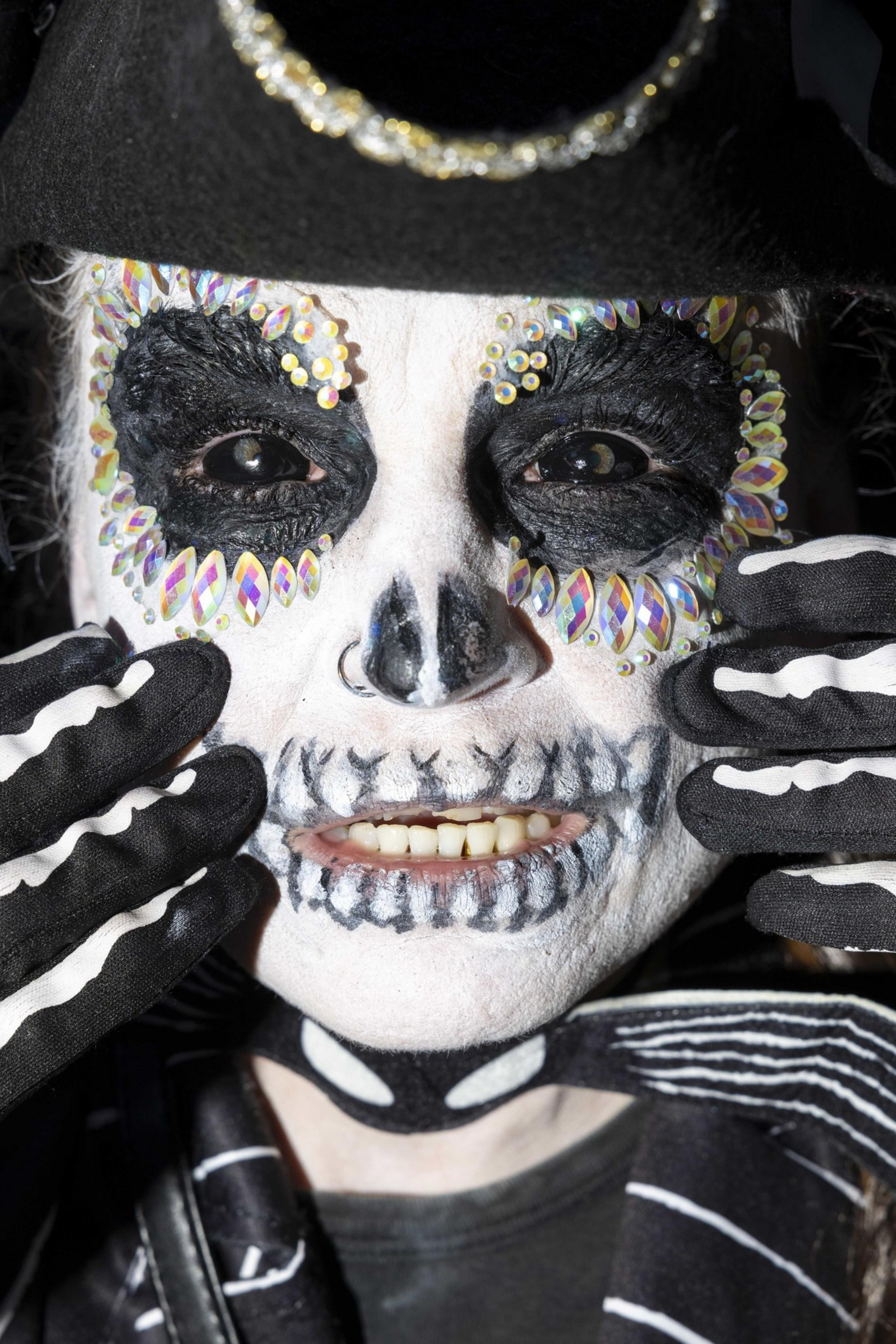 A person with intricate skull face paint, rhinestones around the eyes, and skeleton gloves, wearing a black hat with a glittery band edge.