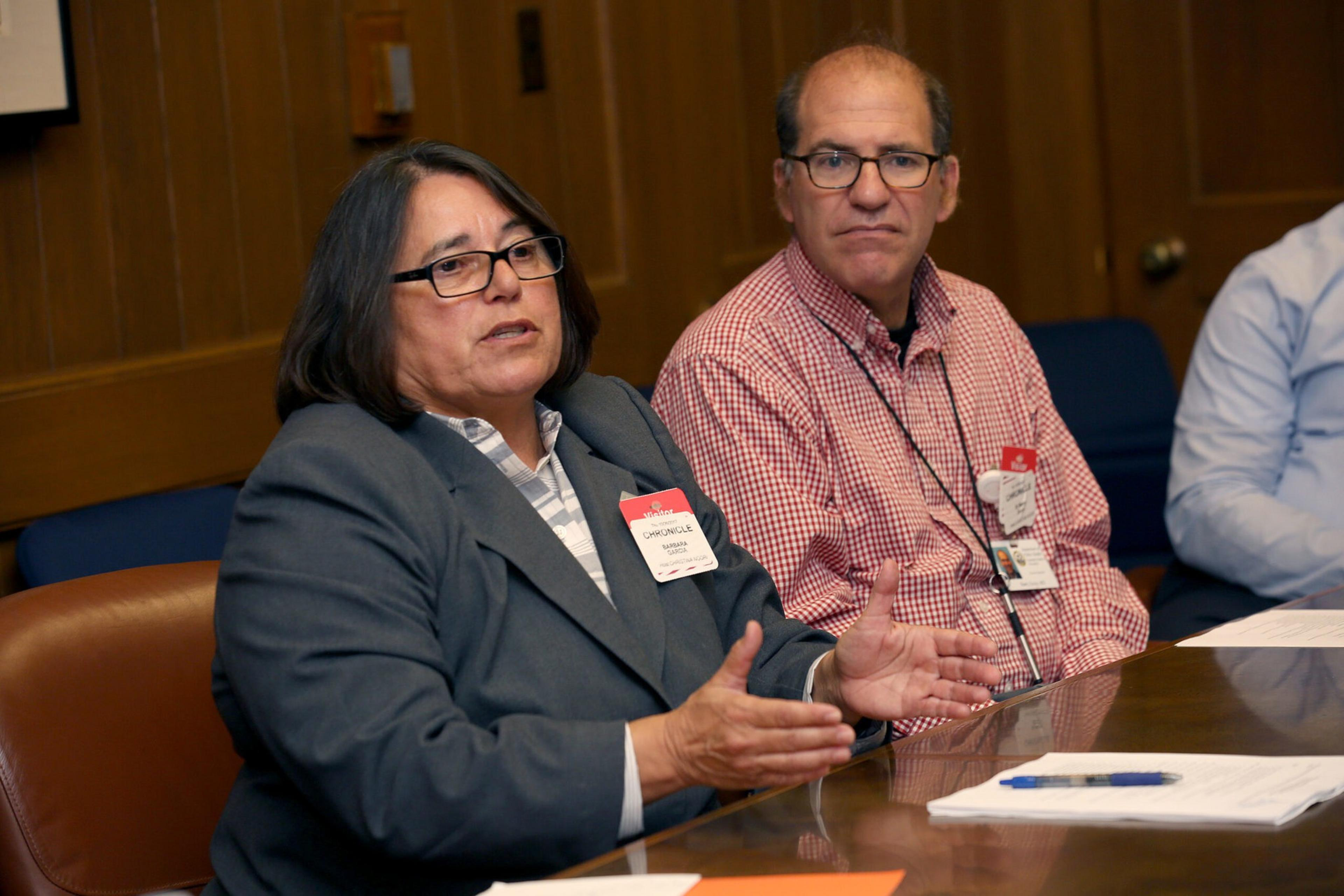 A woman in a gray suit gestures while speaking, seated next to a man in a red checkered shirt listening attentively in a wood-paneled meeting room.