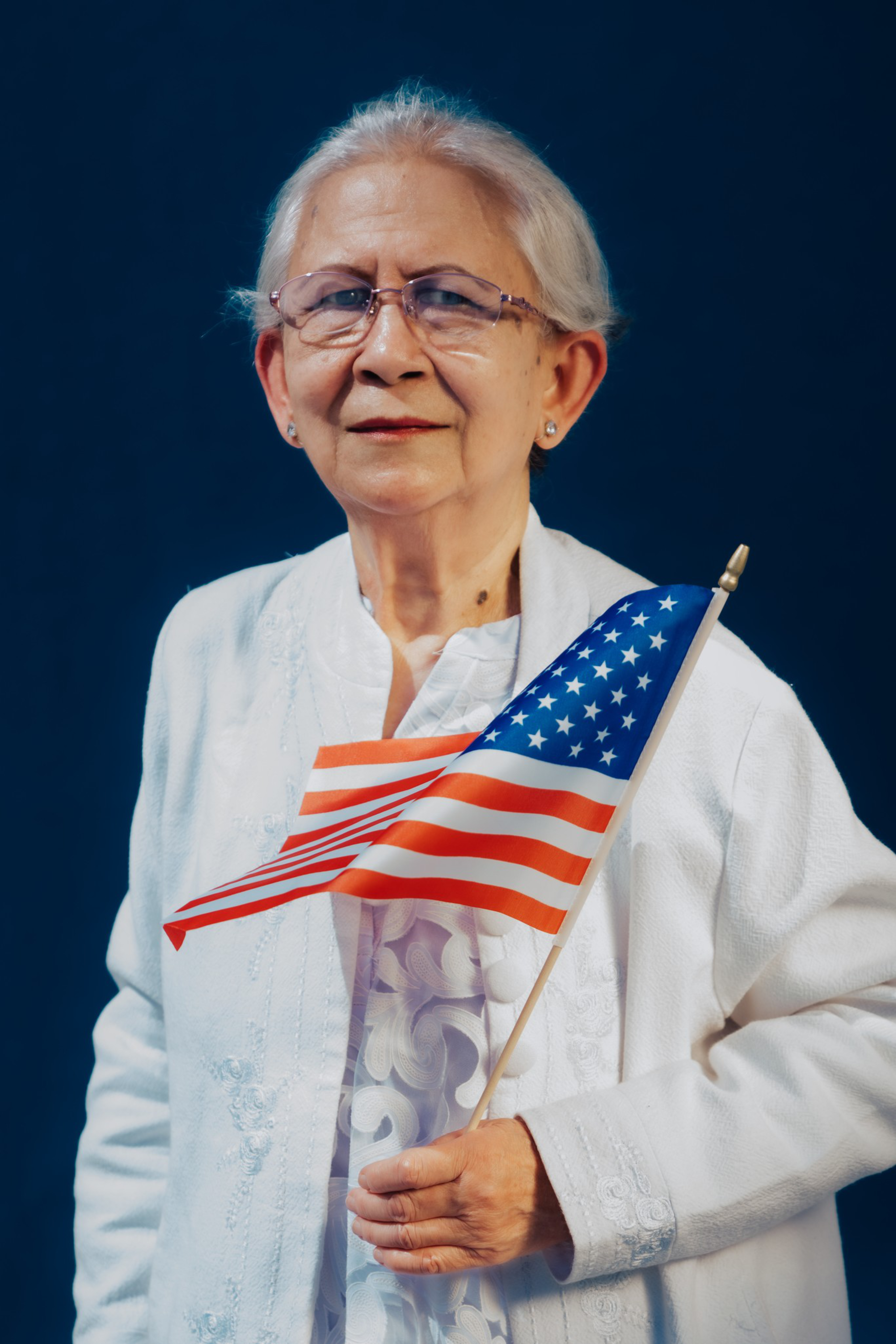 An elderly woman with glasses wears a white embroidered outfit and holds a small American flag against a dark blue background.