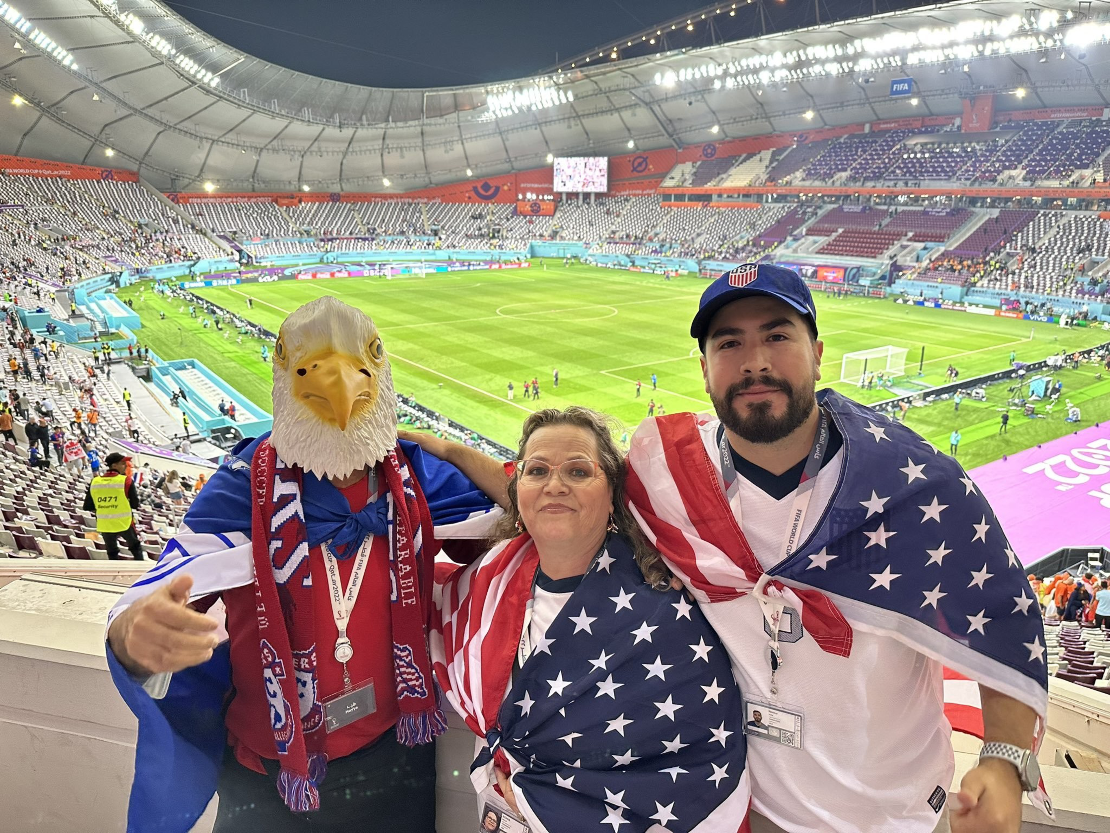Three fans at a soccer stadium wrap themselves in American flags; one wears an eagle mask and all smile with the field and stands behind them.