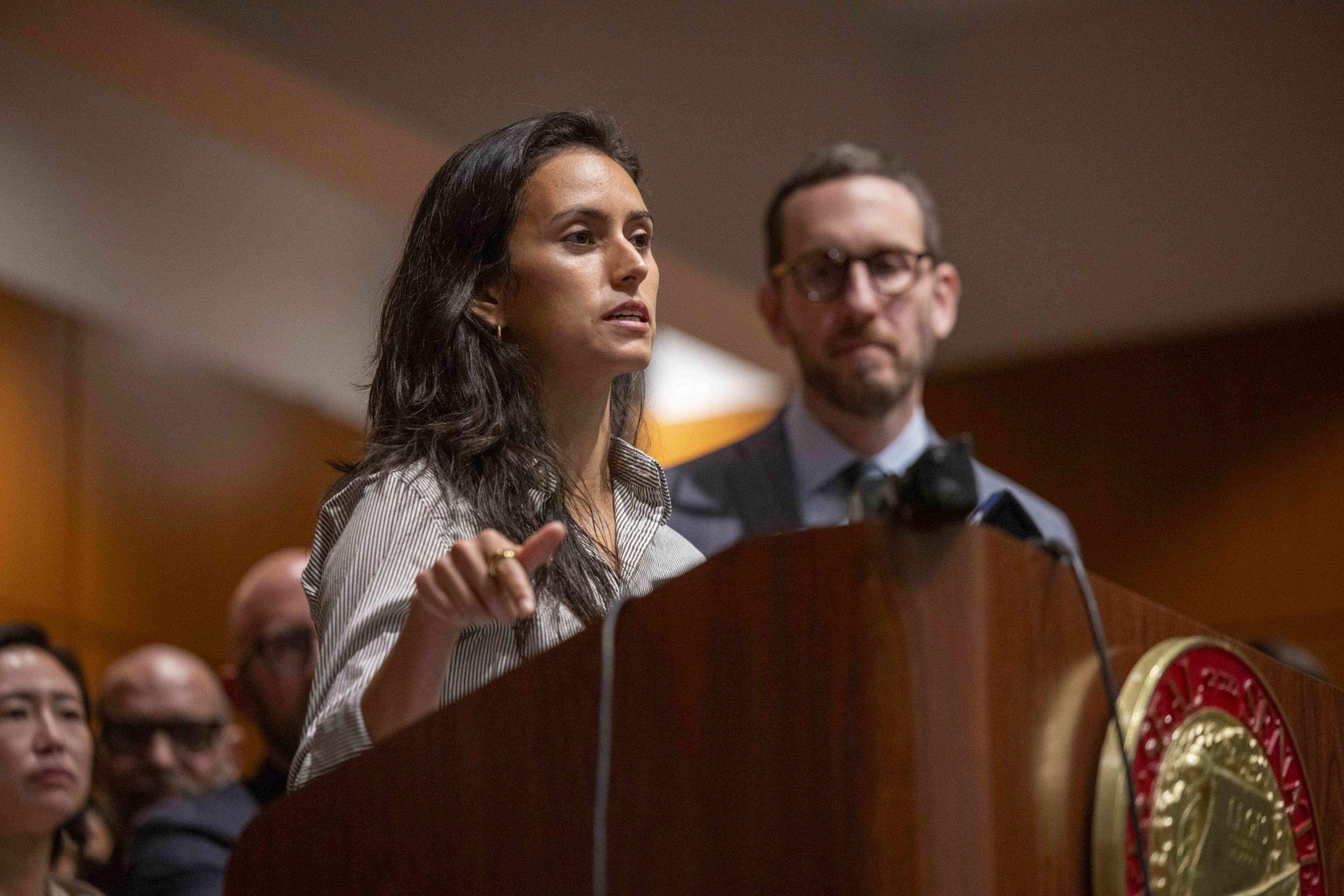 A woman speaks passionately at a podium, pointing, with a man standing behind her. The scene appears to be a formal event, with several people in the background.