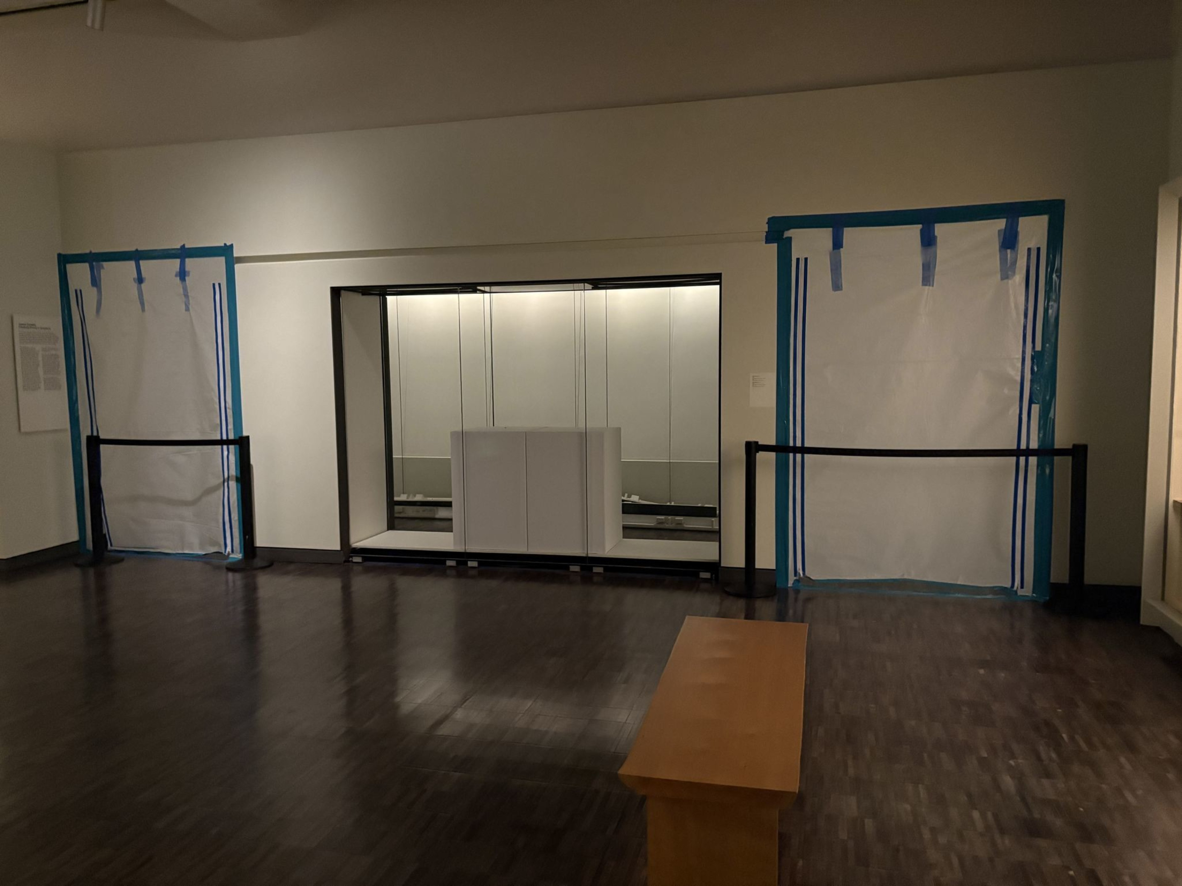 The image shows an empty room with reflective flooring, a wooden bench, and two blue-taped panels covering sections of the walls. A glass display case is in the center.