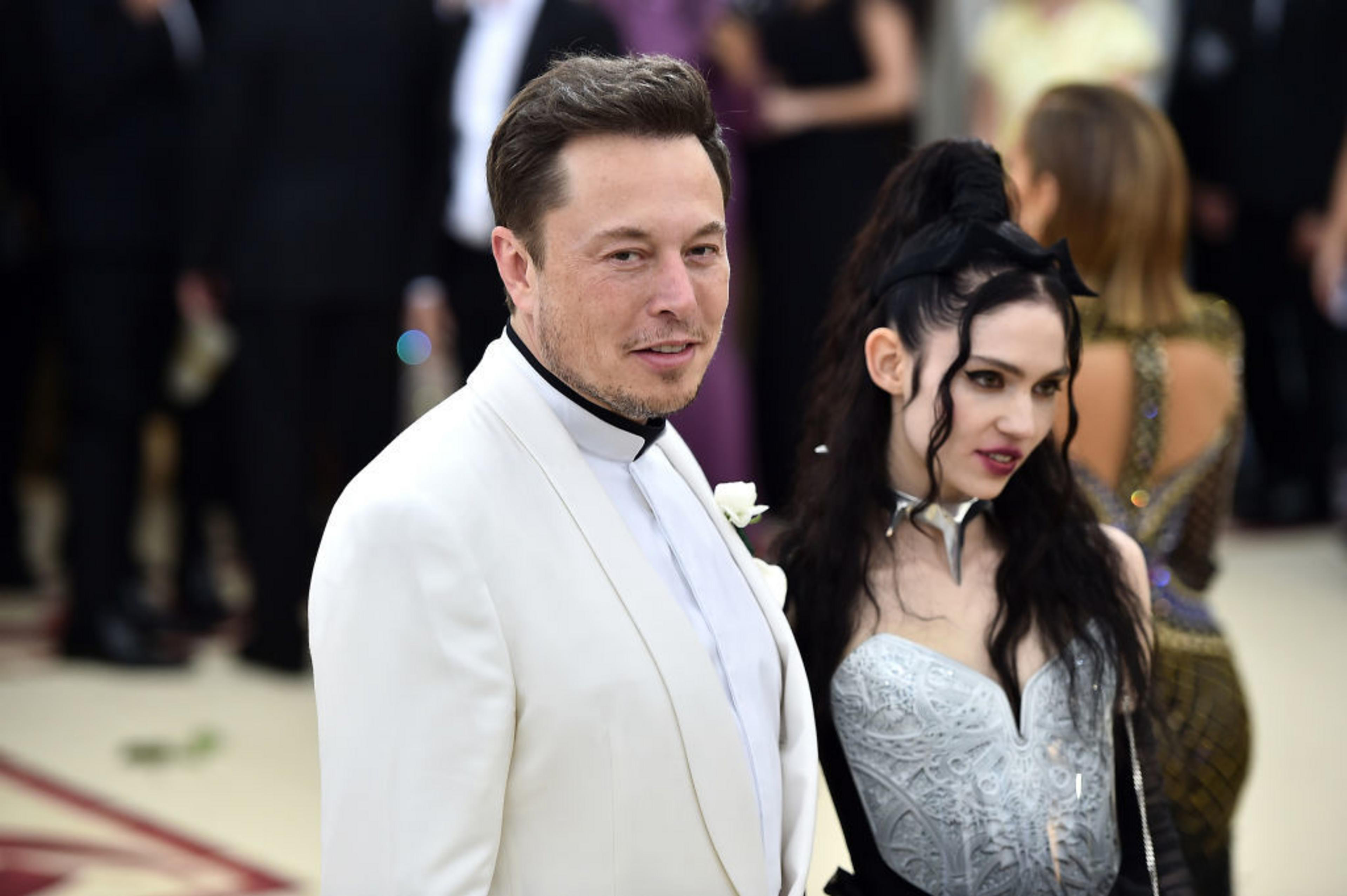 Elon Musk, wearing a white suit and Grimes, wearing a silver dress, are seen from the side standing close together in a photo showing them attending the Heavenly Bodies: Fashion and the Catholic Imagination Costume Institute Gala at The Metropolitan Museum
