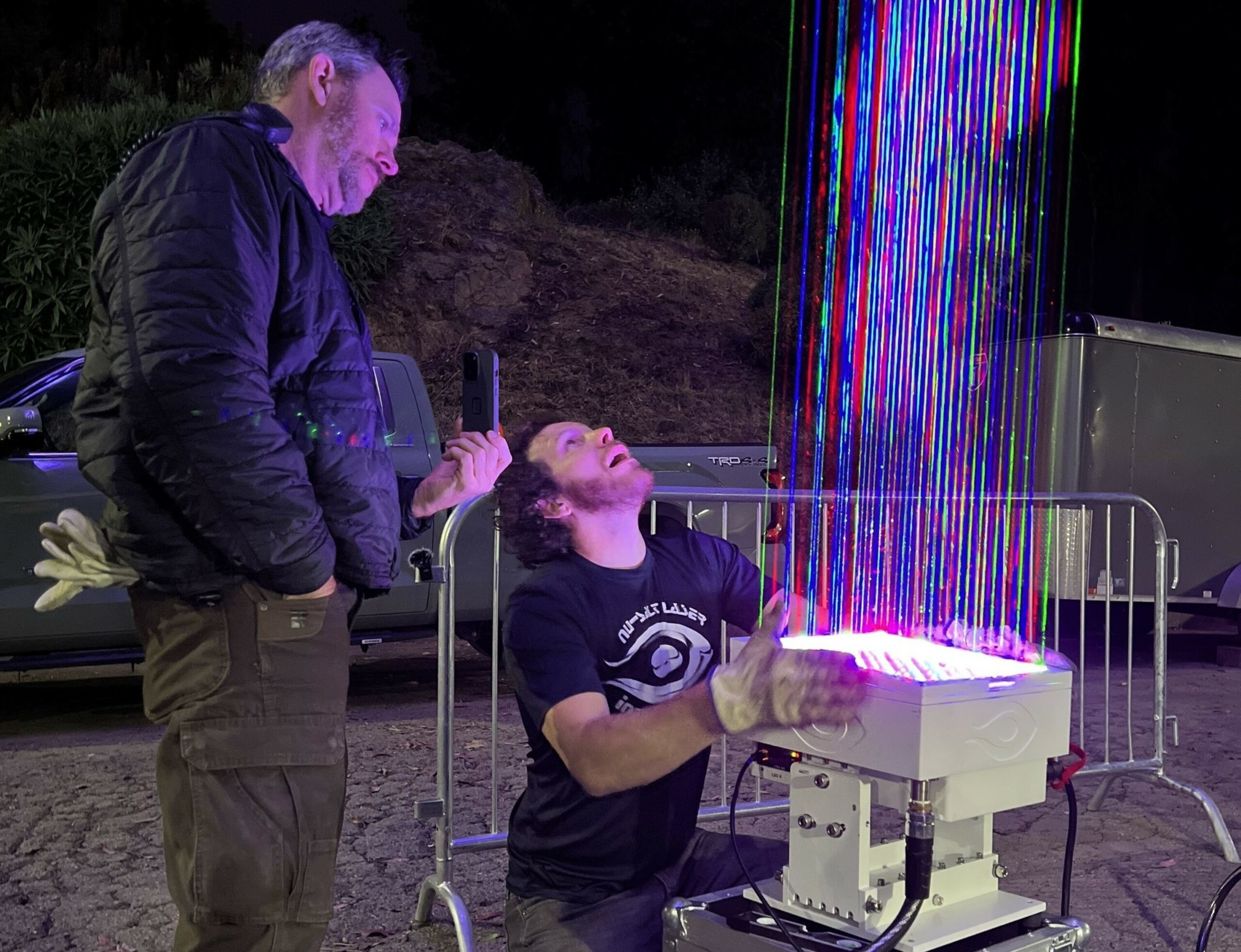 A man kneeling with gloves reaches toward a device emitting vertical, multicolored light beams while another man nearby watches and records with a phone.