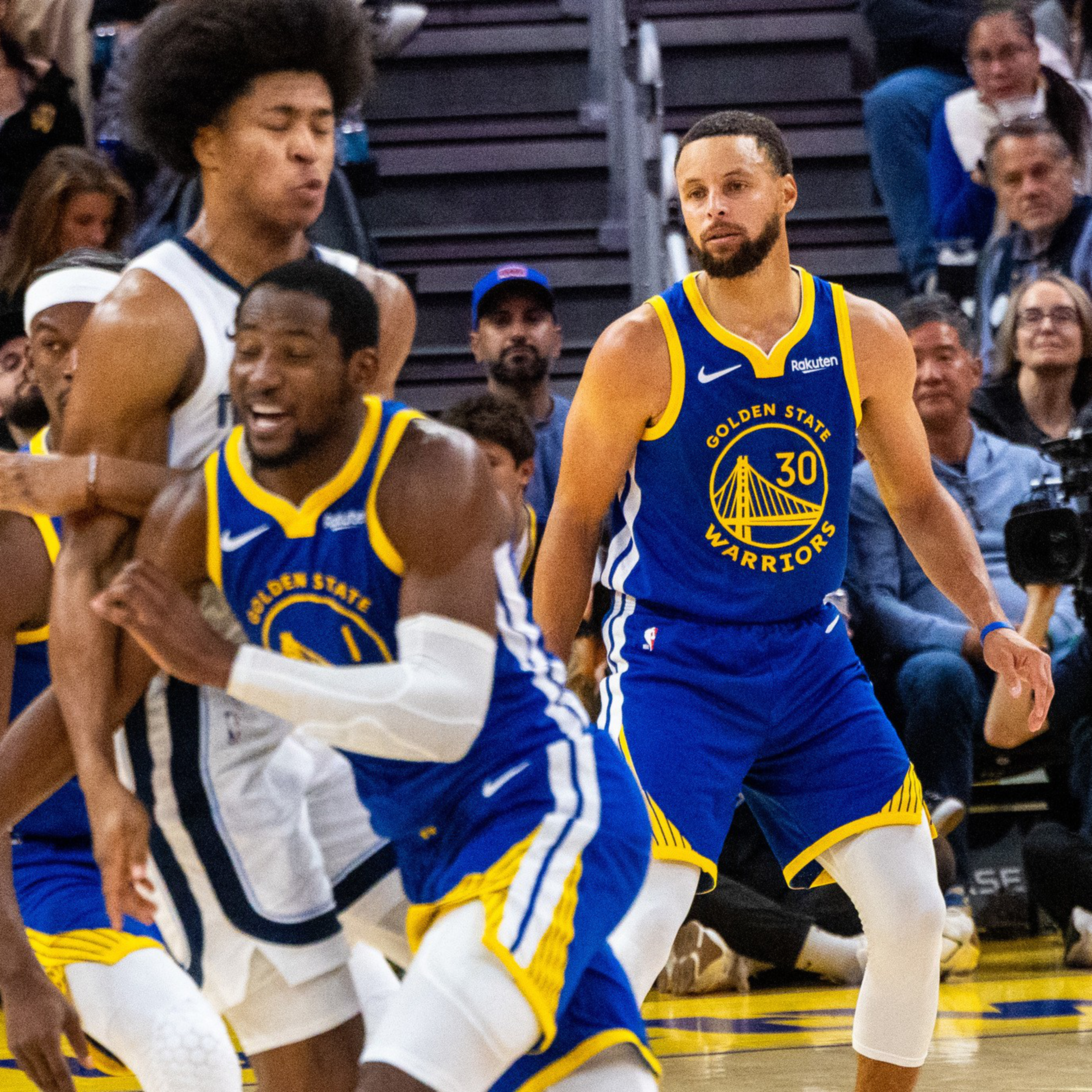 Two Golden State Warriors players in blue uniforms move defensively against an opposing player in white during a basketball game, with spectators watching.