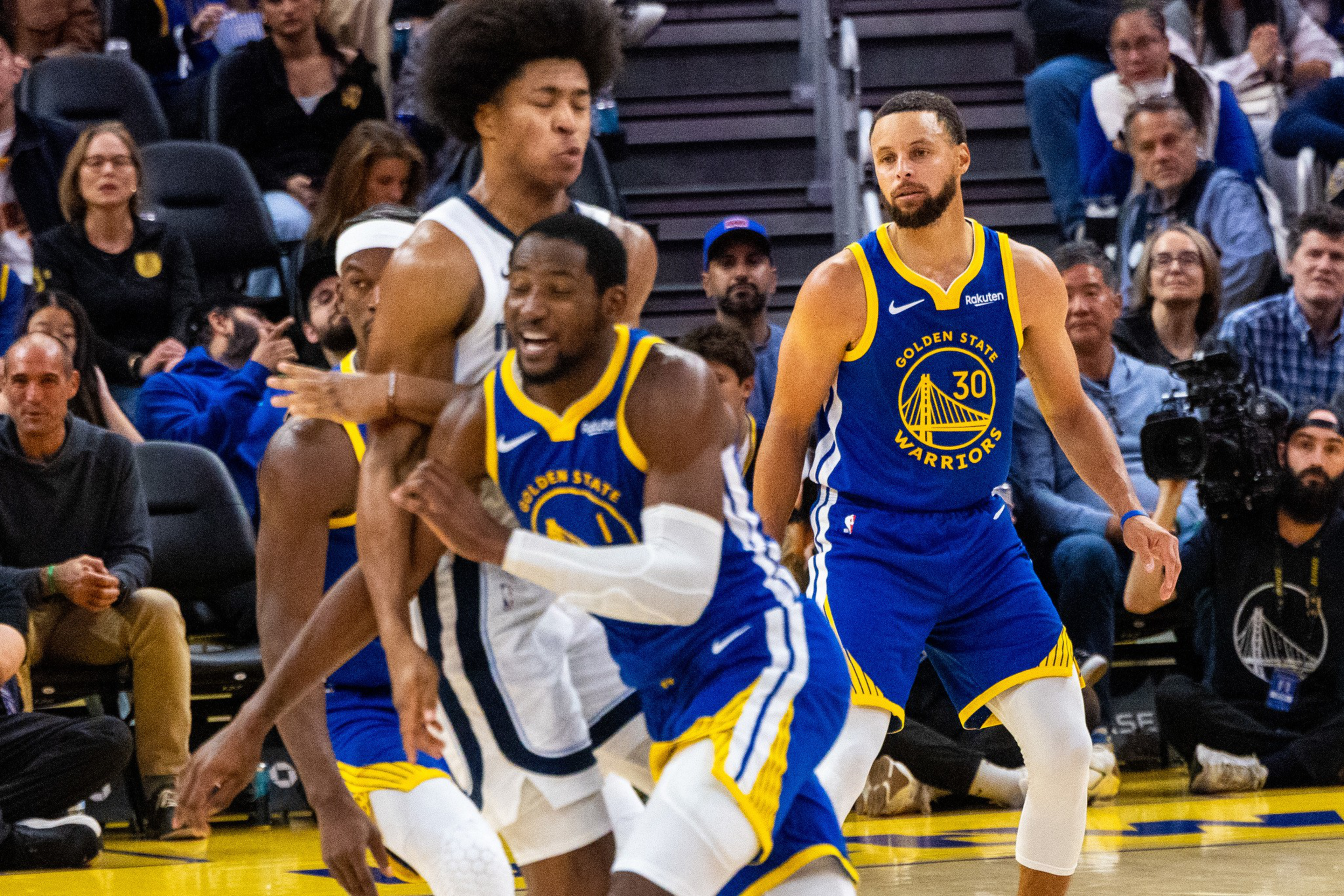 Two Golden State Warriors players in blue uniforms move defensively against an opposing player in white during a basketball game, with spectators watching.
