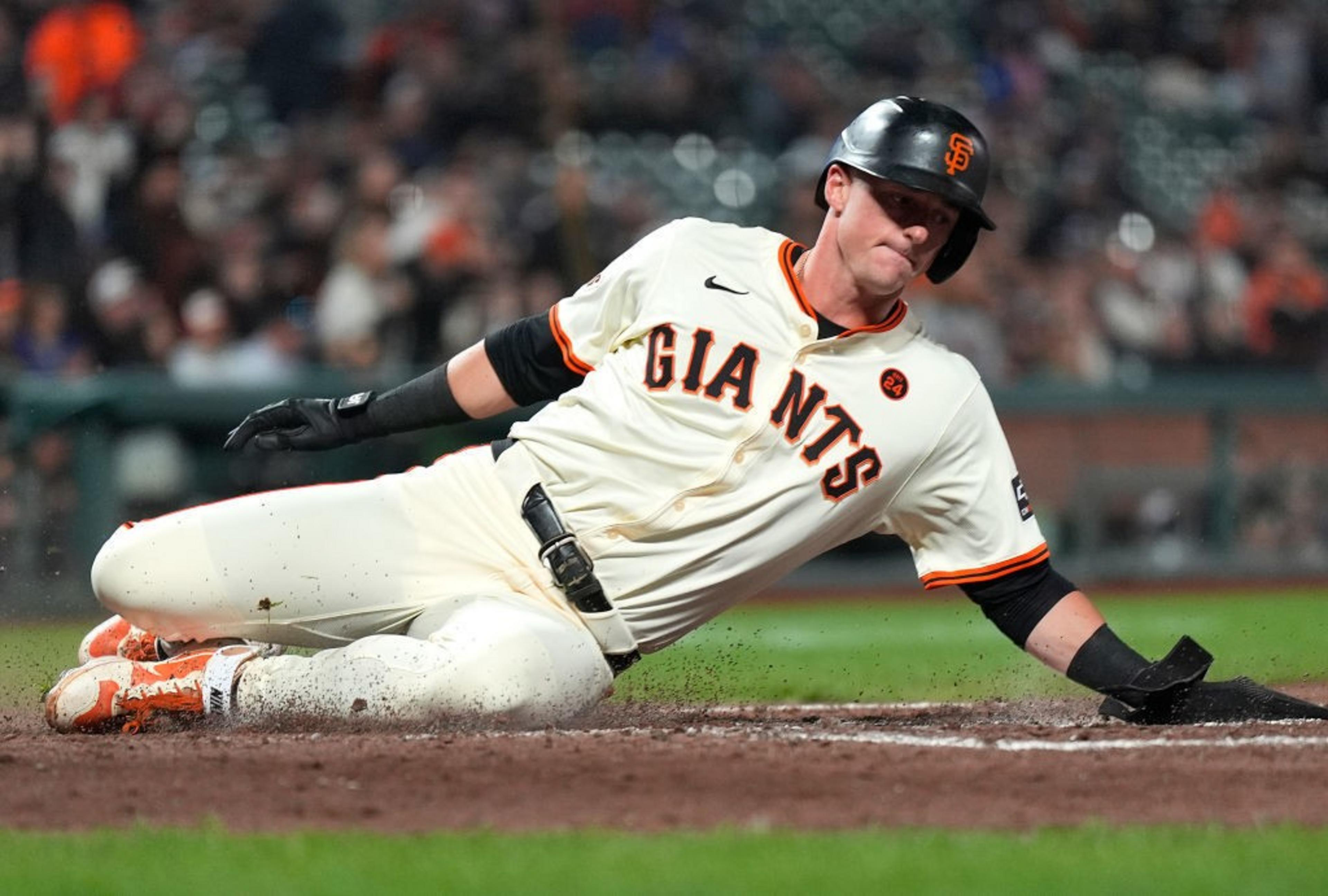 A baseball player wearing a white "Giants" uniform and helmet slides into a base on a dirt field, with a focused expression during a game.