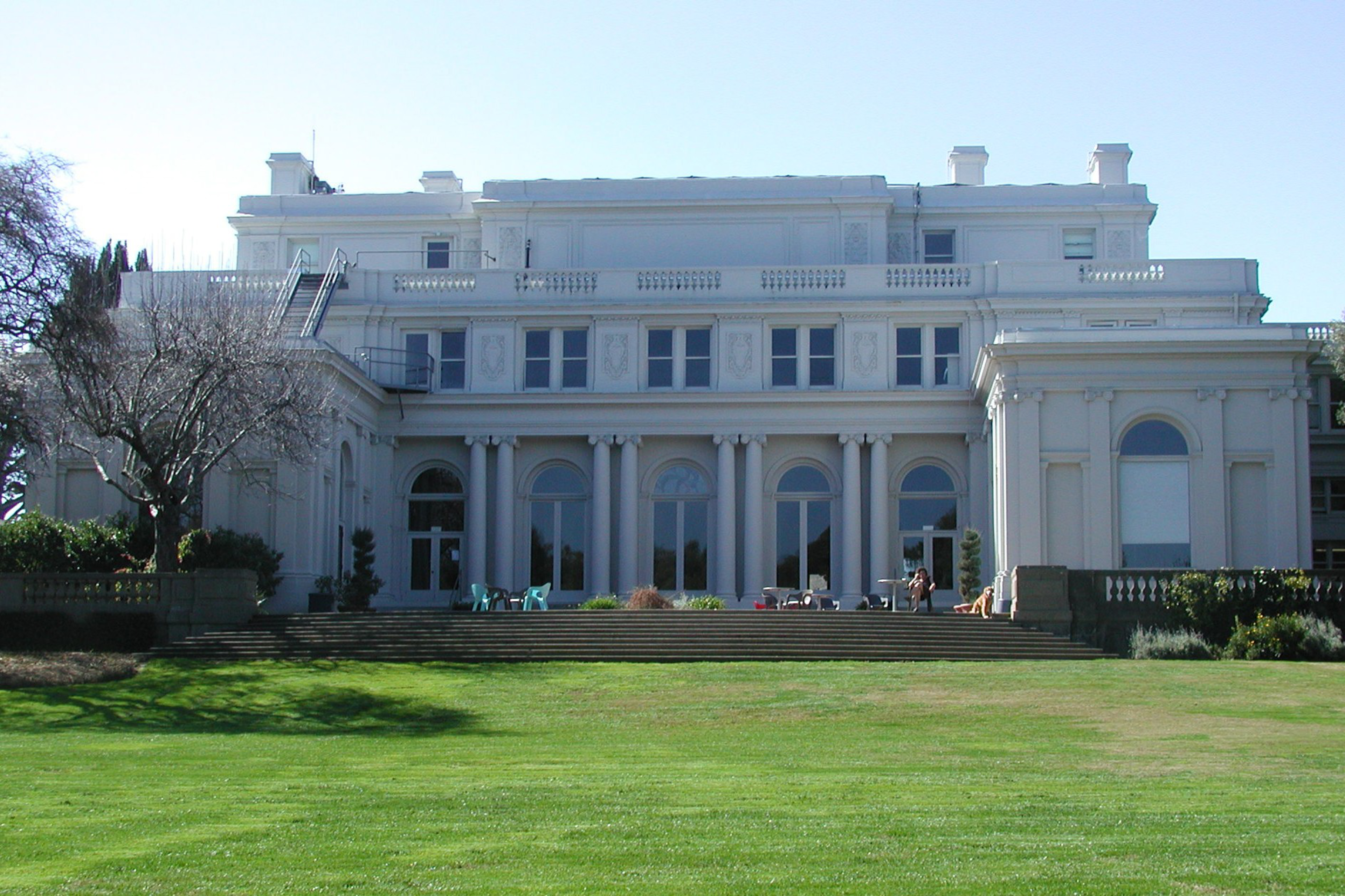 A large white neoclassical mansion with tall columns, arched windows, and a lawn in front under a clear blue sky.