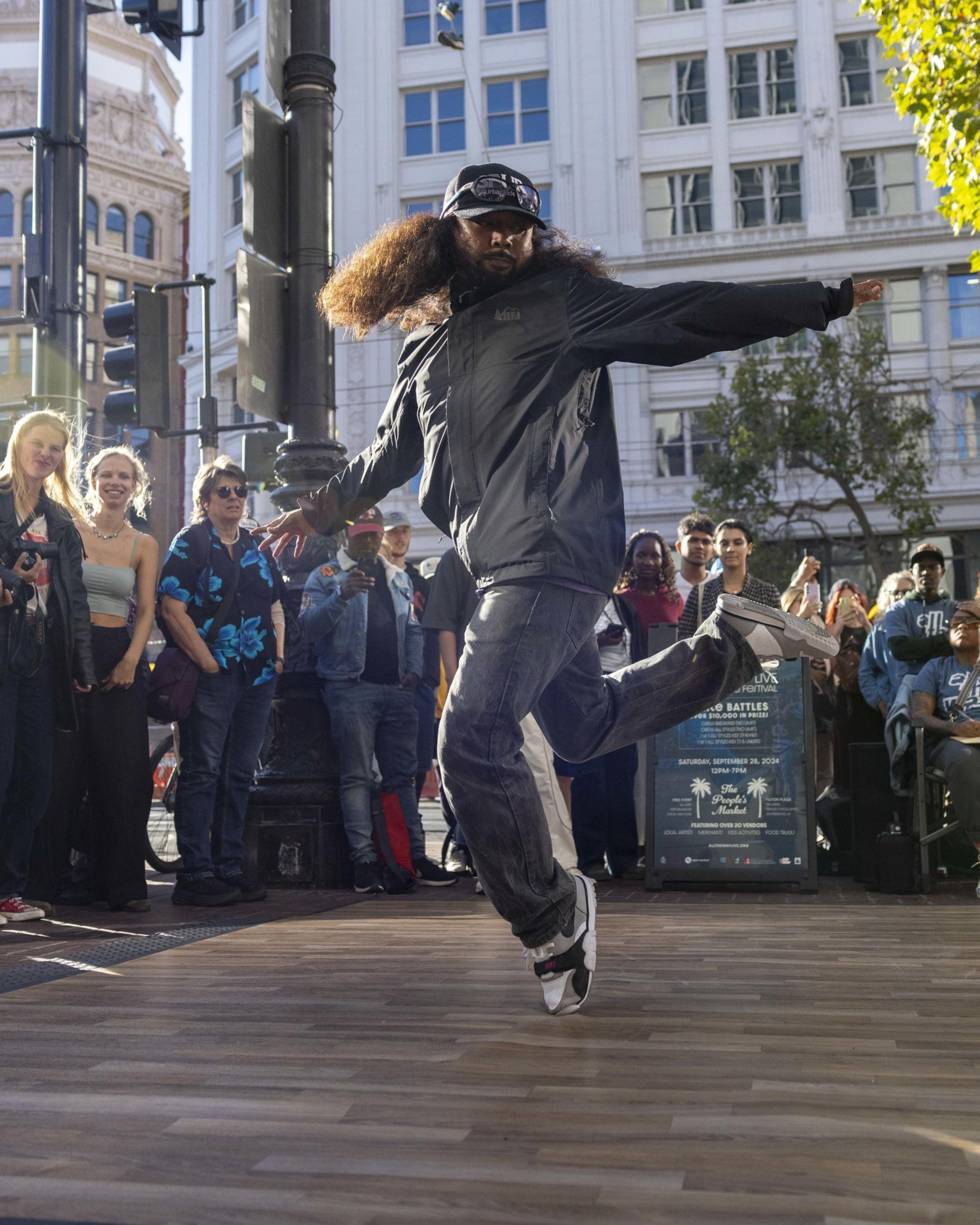 A street dancer with thick, curly hair performs energetically in front of an engaged crowd on a wooden platform in an urban area with tall buildings and trees.