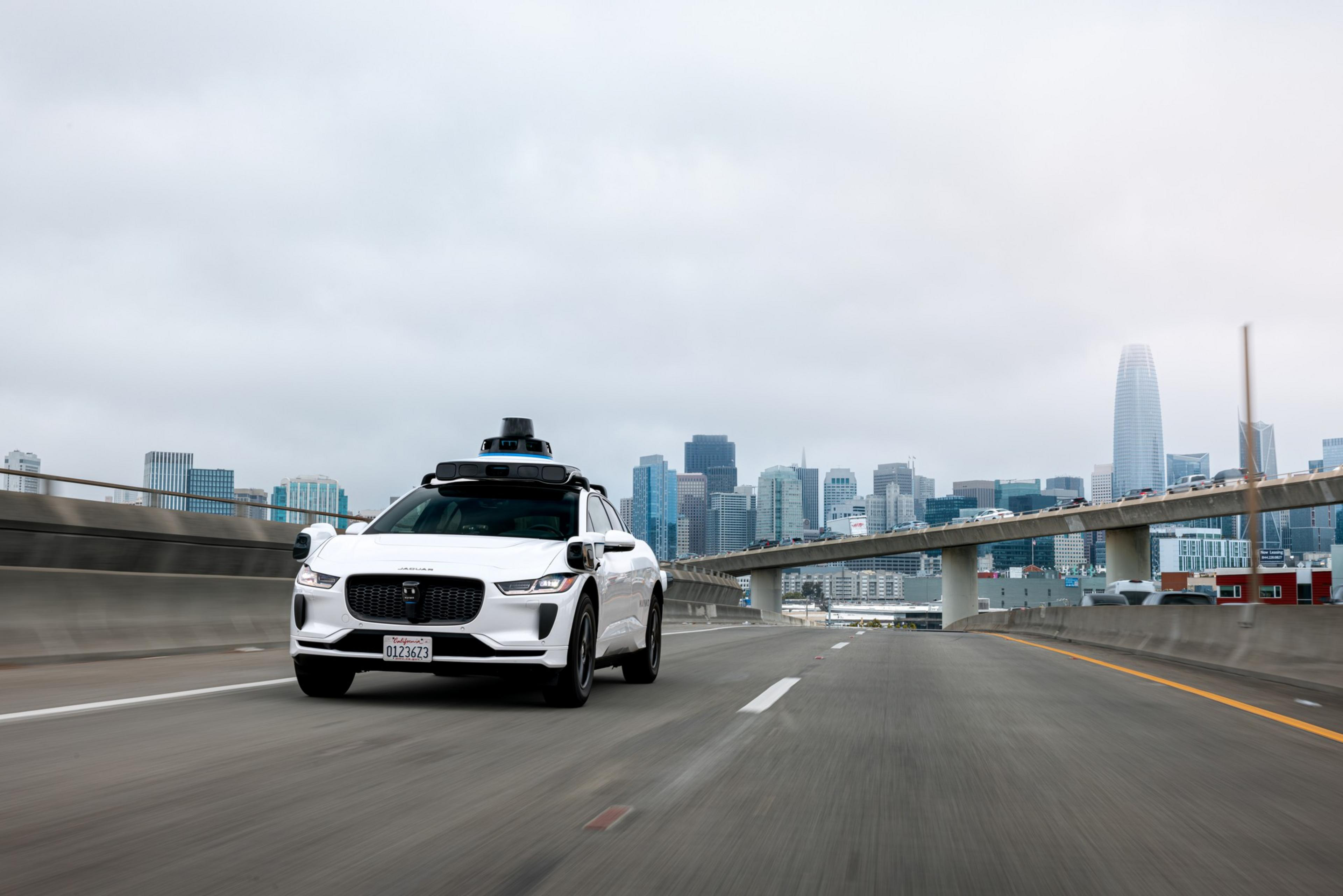 A white self-driving car equipped with sensors drives on an empty highway with a city skyline and an overpass in the background under a cloudy sky.