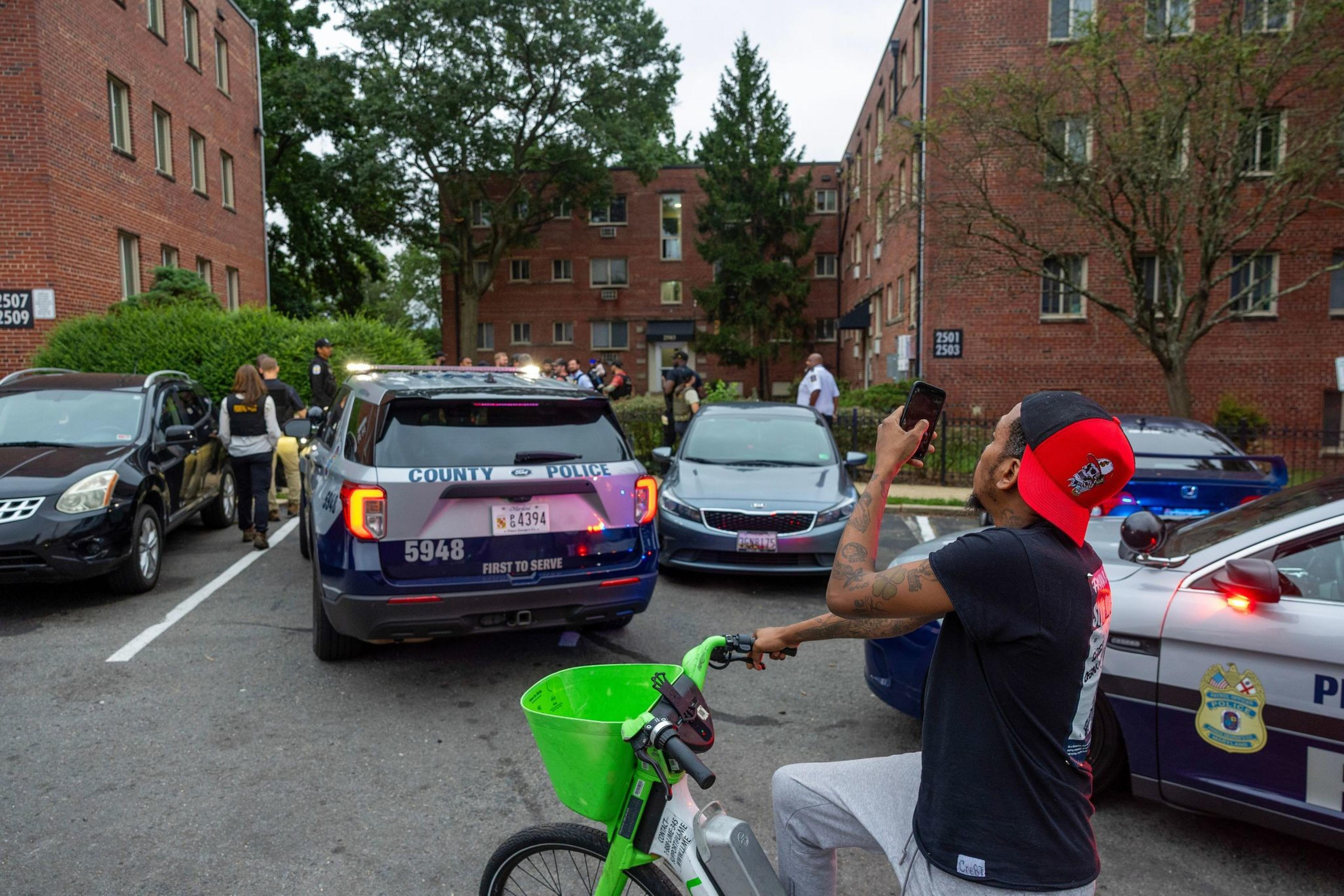 A man on a green bike films a police scene where officers talk to people near red brick apartment buildings and parked cars.