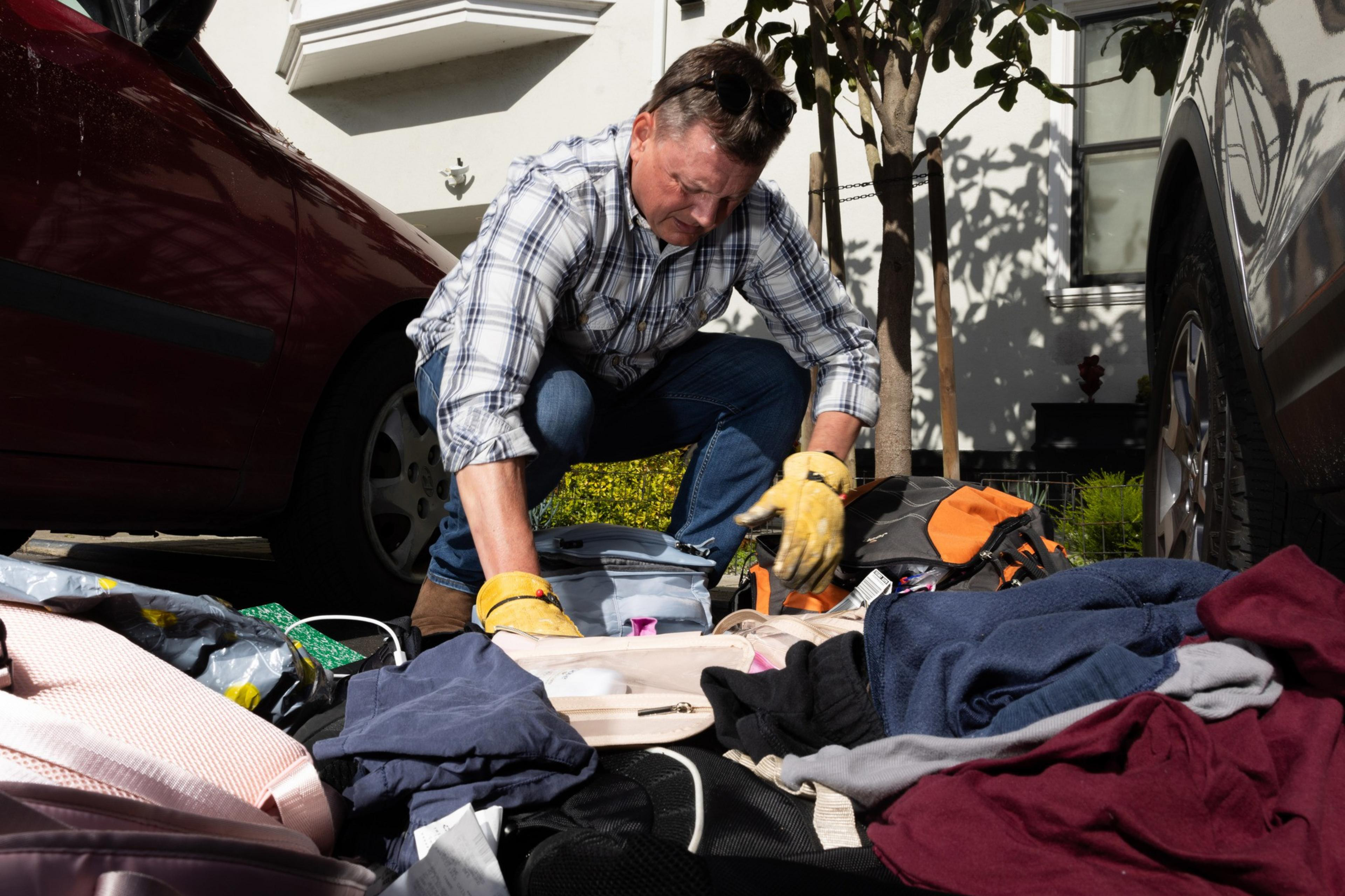 A man kneels outdoors, sorting through assorted items on the ground between two parked cars, wearing gloves and a focused expression.
