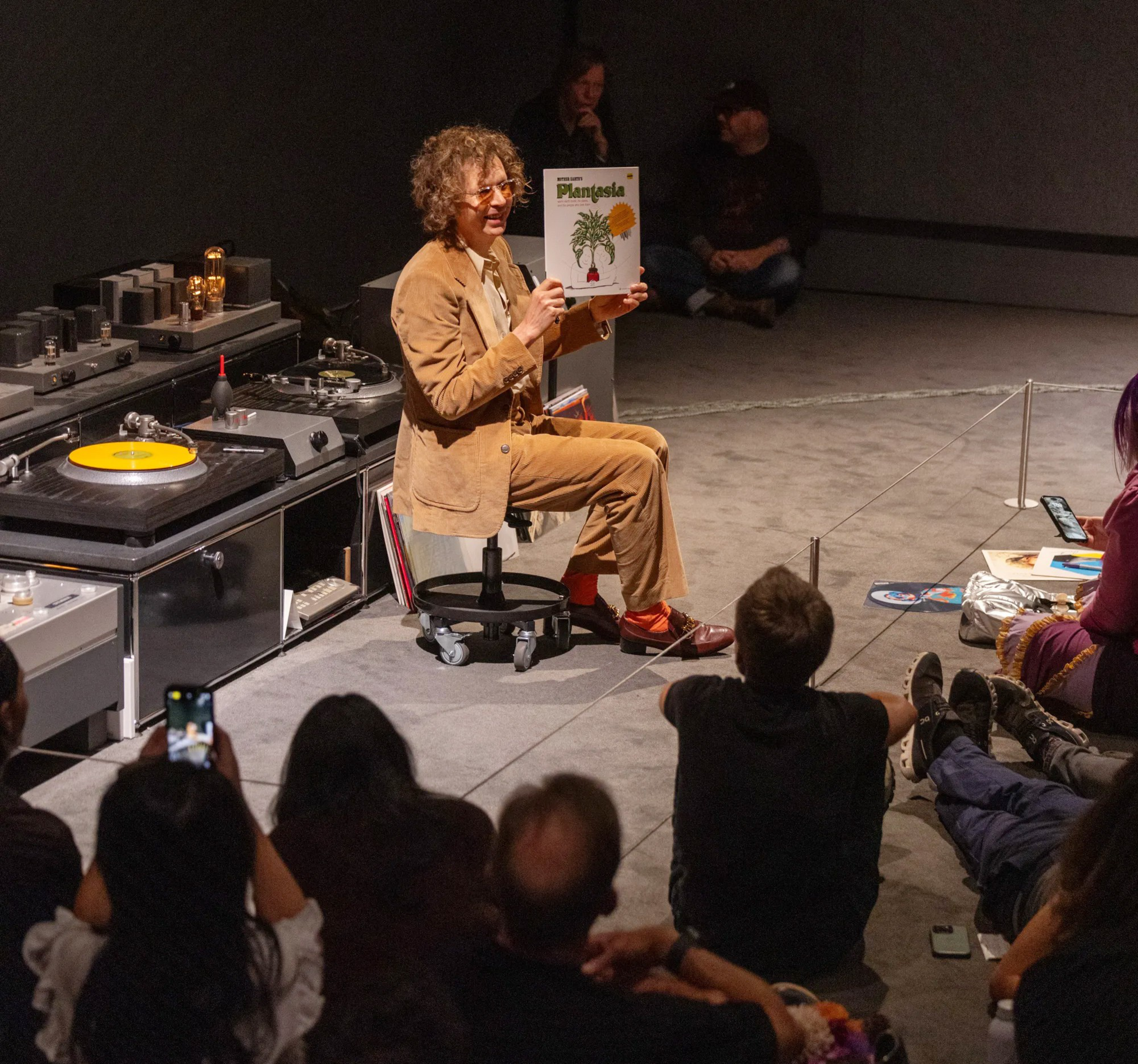 A person in a brown suit is sitting on a stool, holding a record cover titled &quot;Plantasia.&quot; They are surrounded by an audience seated on the floor, watching attentively.