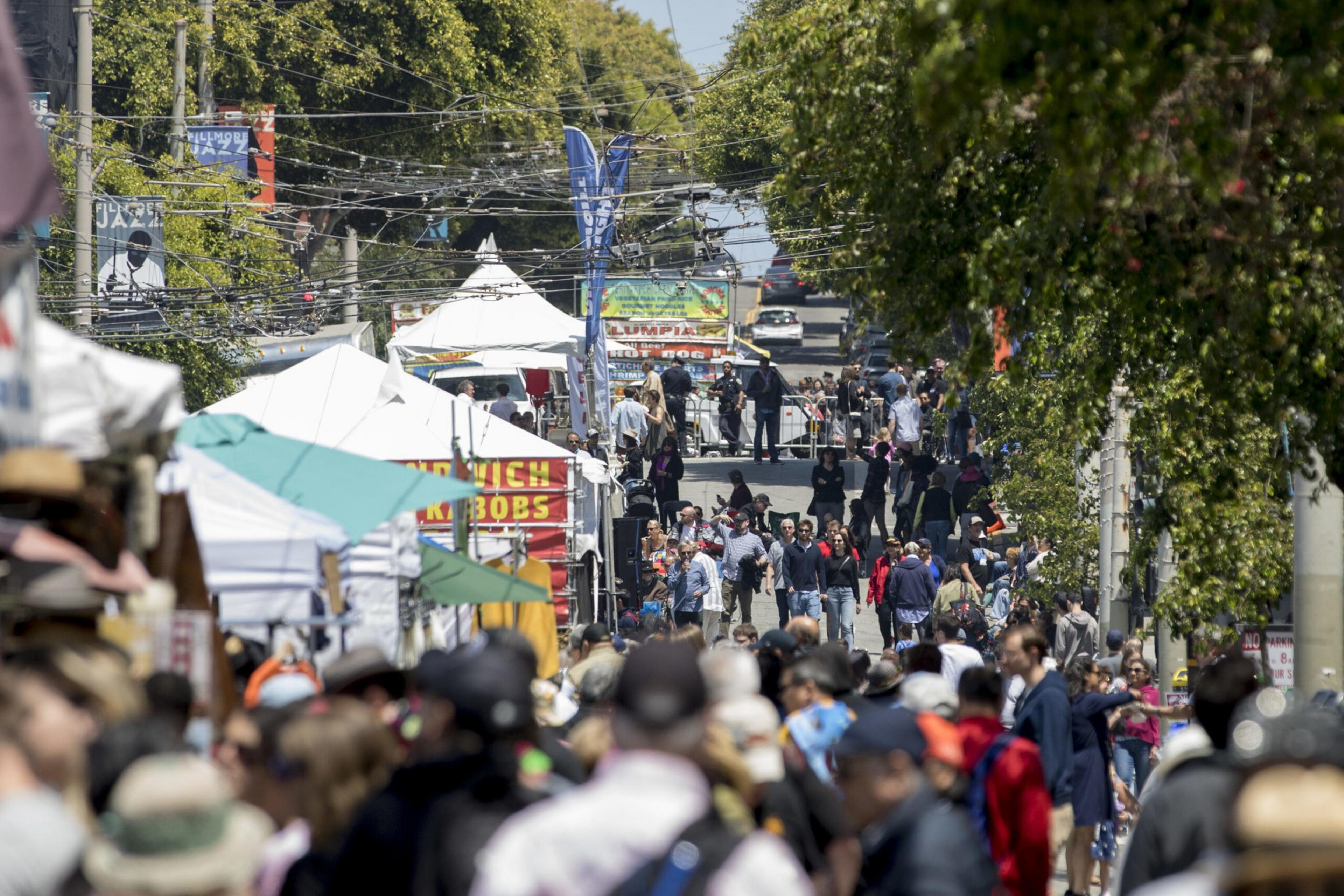 A bustling street market is filled with people walking among food stalls and tents, surrounded by trees and colorful banners overhead.