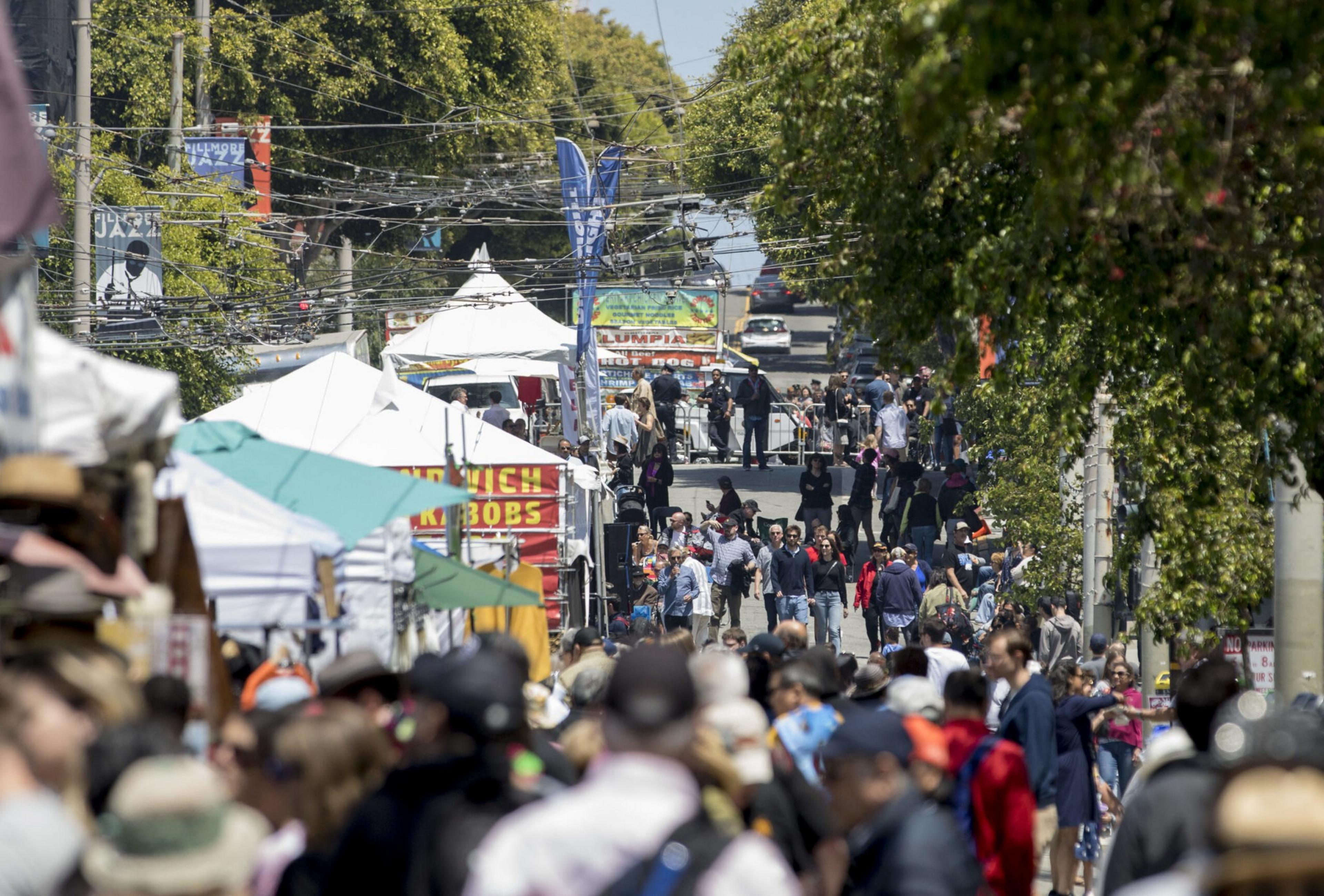 A bustling street market is filled with people walking among food stalls and tents, surrounded by trees and colorful banners overhead.