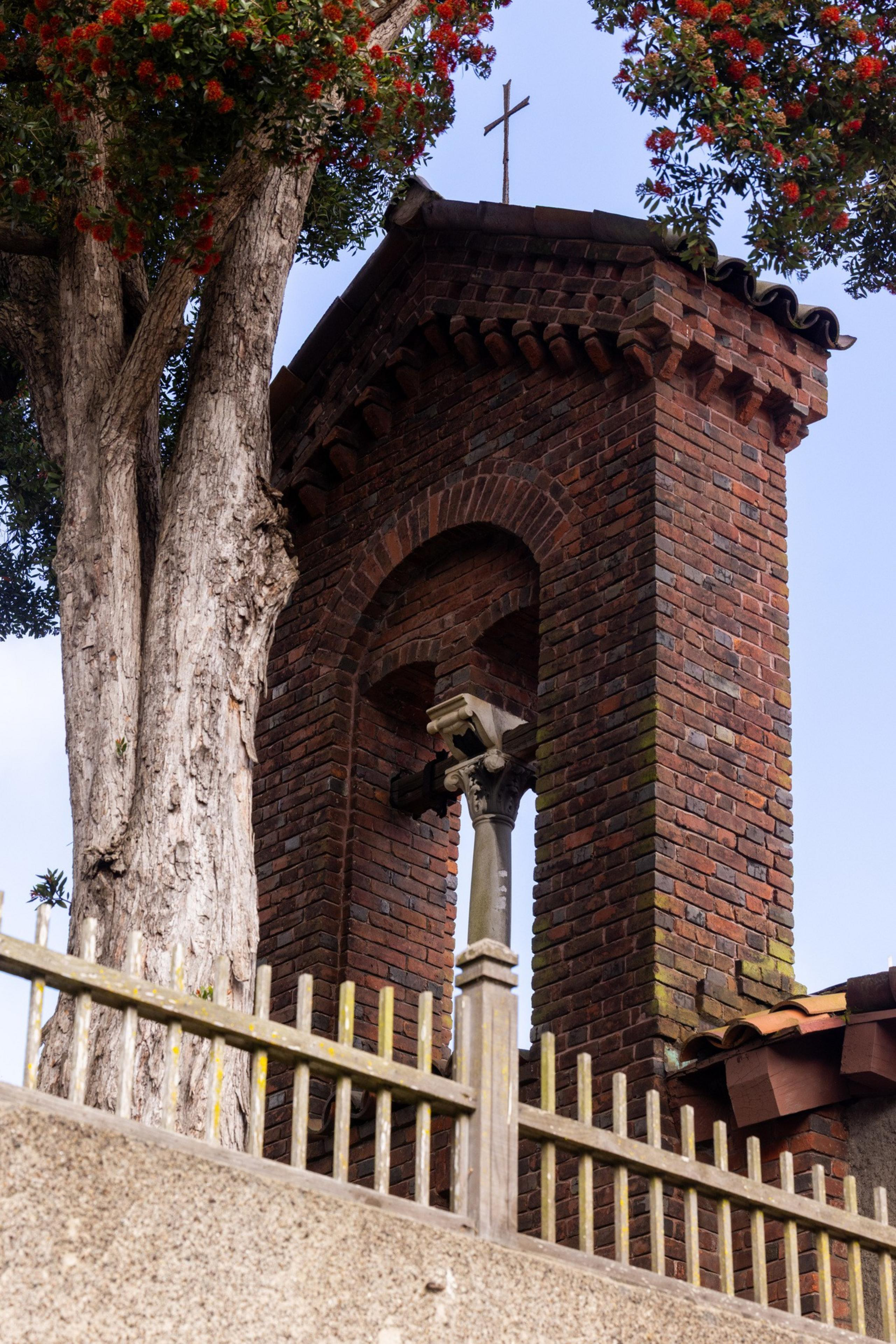 A brick church tower with a cross on top is framed by a tall tree with red flowers. A wooden fence is visible in the foreground.