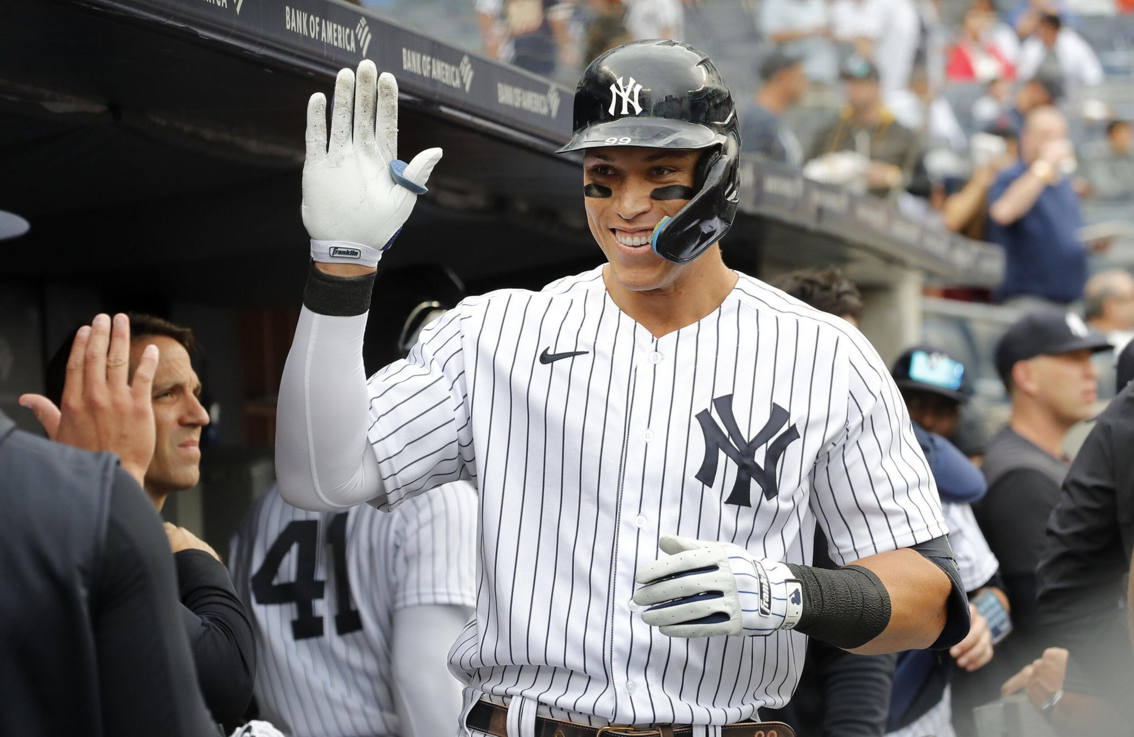 A smiling New York Yankees player wearing a helmet and striped uniform raises his gloved hand for a high-five near the dugout.