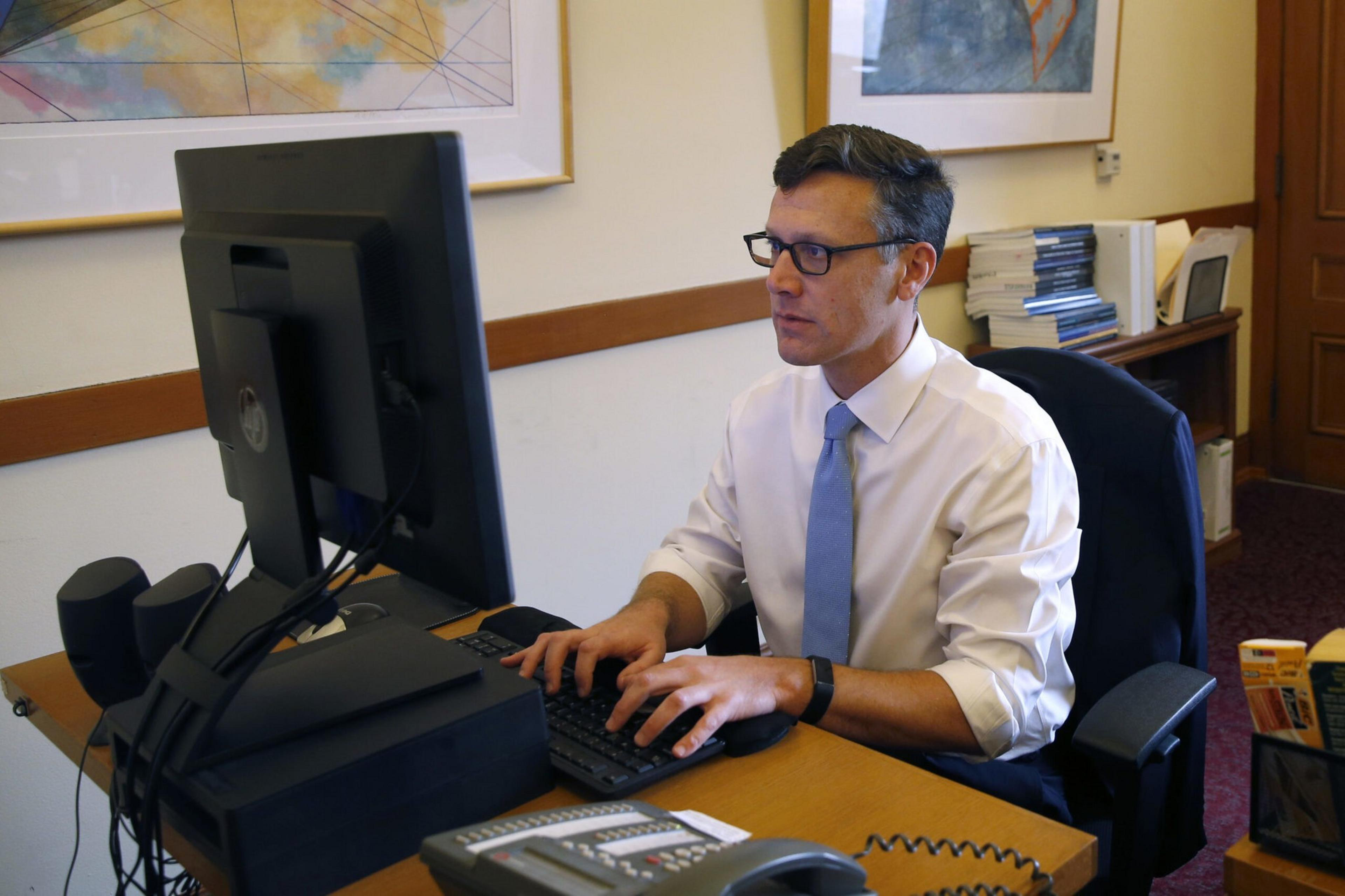A man in a white shirt and blue tie types on a keyboard at a desk with a computer, phone, and stacks of papers in an office.