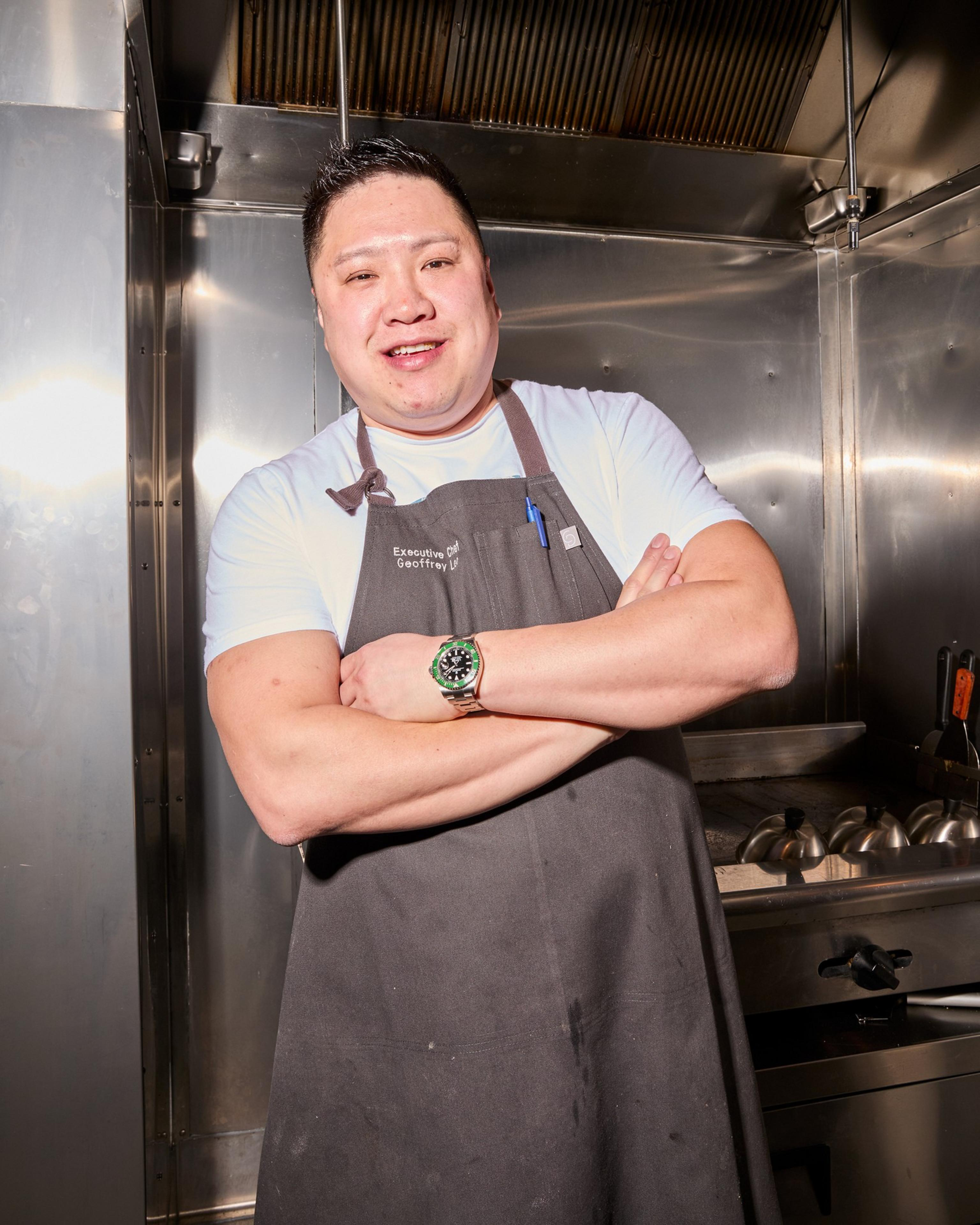 A person stands in a kitchen, smiling confidently with arms crossed. They're wearing a dark apron over a white shirt and a watch, with a pen in the apron pocket.