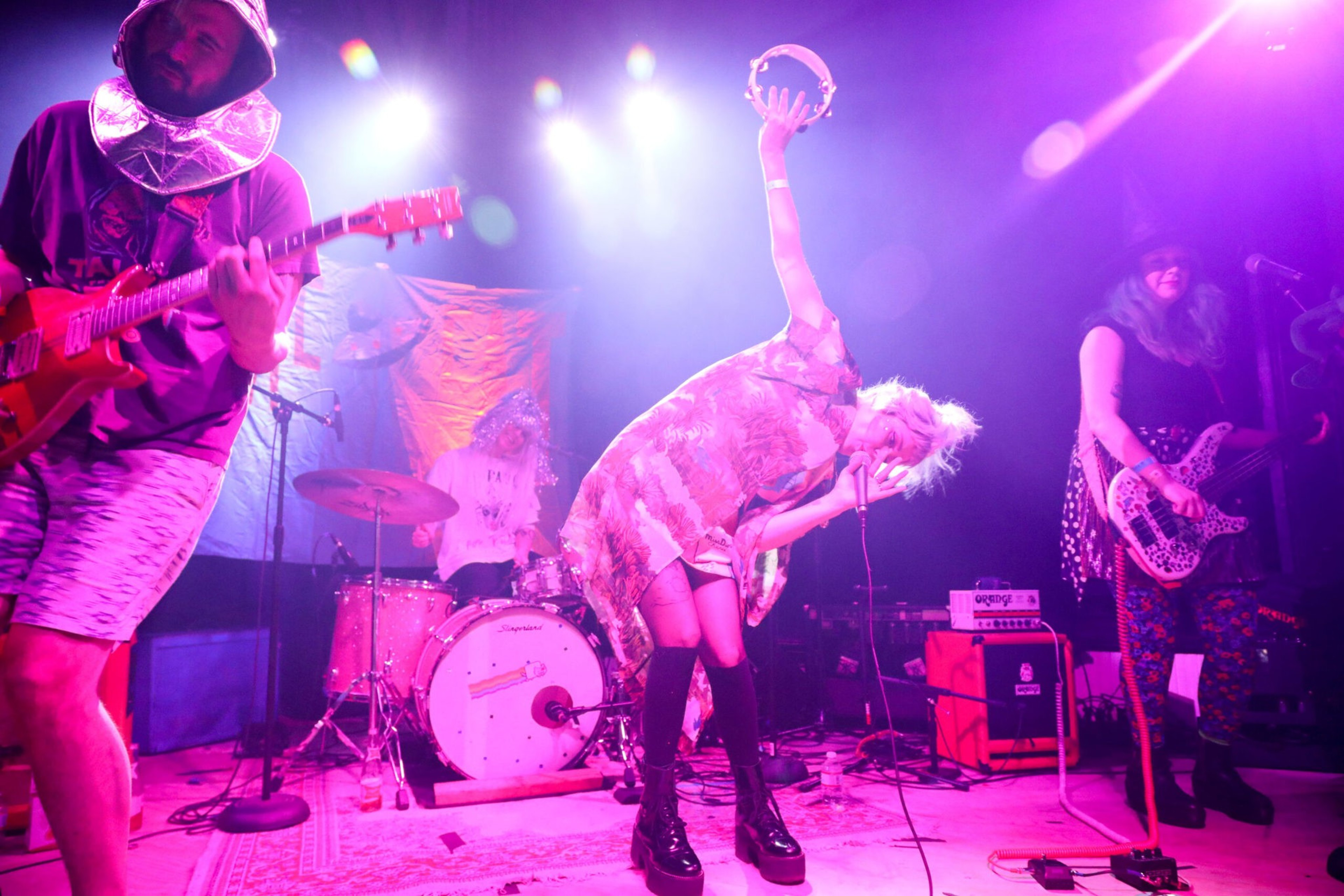 A band performs on stage with colorful lights; the singer raises a tambourine while leaning forward, surrounded by guitarists and a drummer.