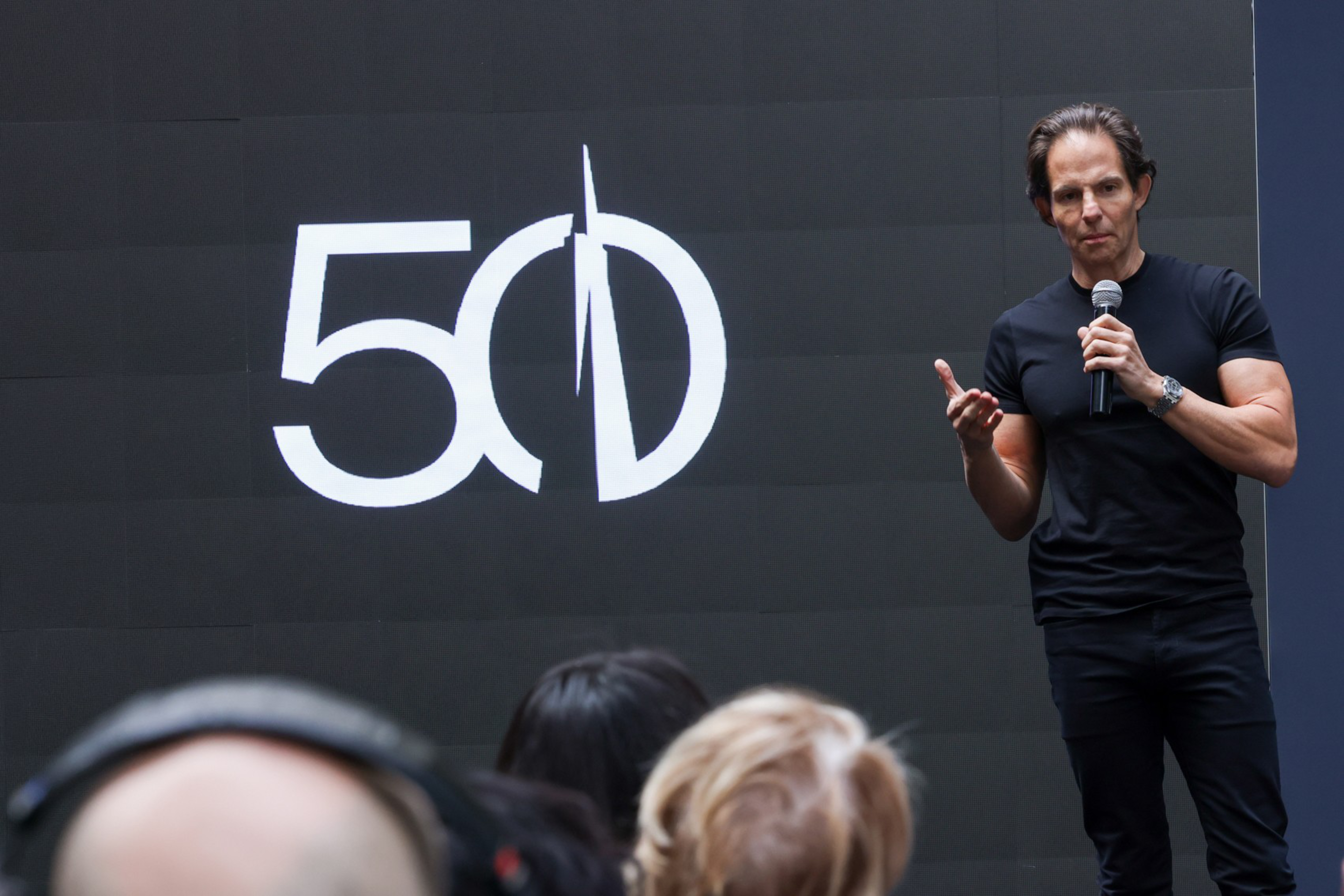A man in a black shirt is speaking into a microphone on a stage with a black background displaying a large white &quot;50&quot; and stylized &quot;D&quot; behind him.