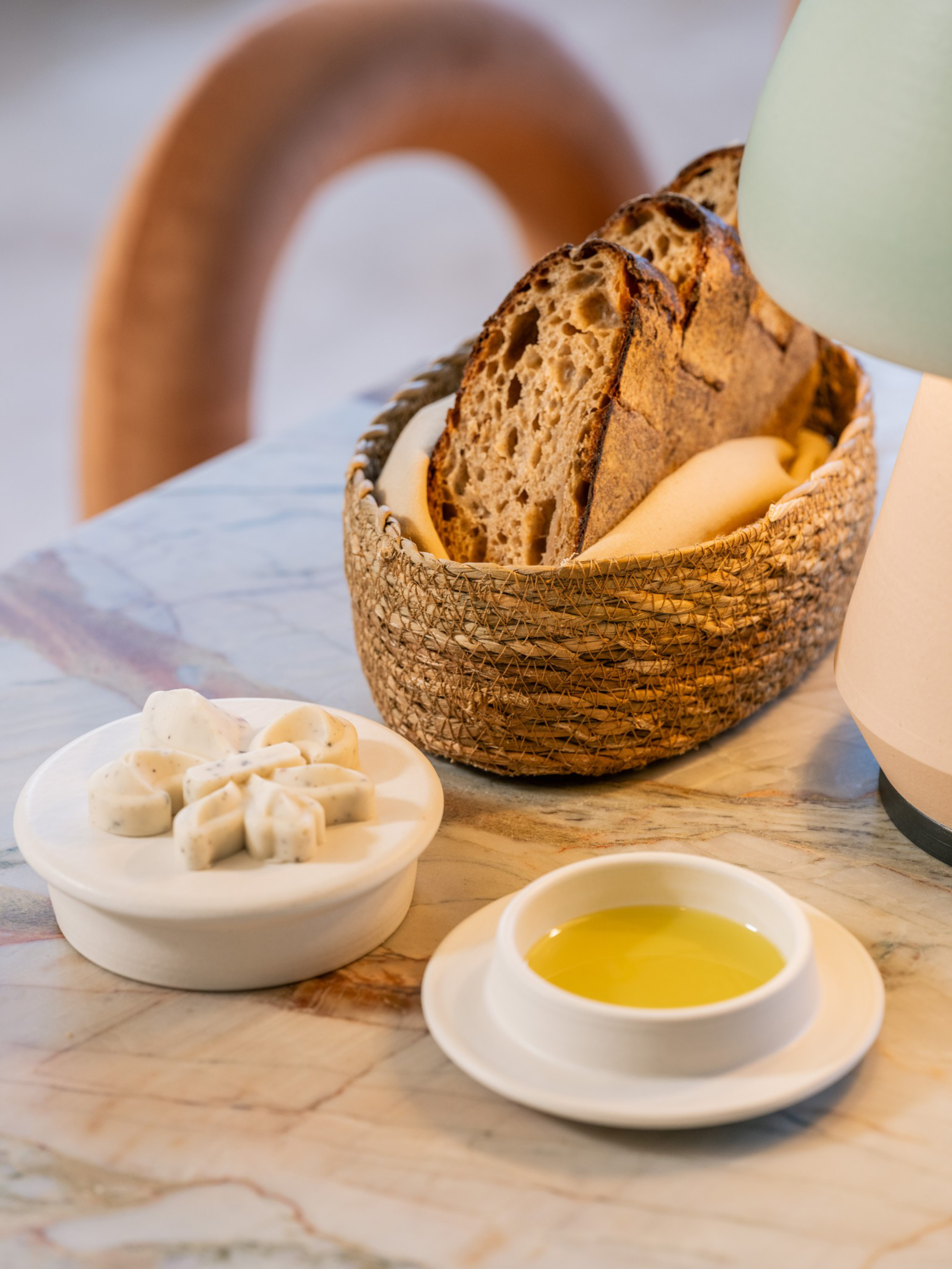 Slices of crusty bread rest in a woven basket lined with cloth, accompanied by a dish of herb butter and a small bowl of olive oil on a marble table.