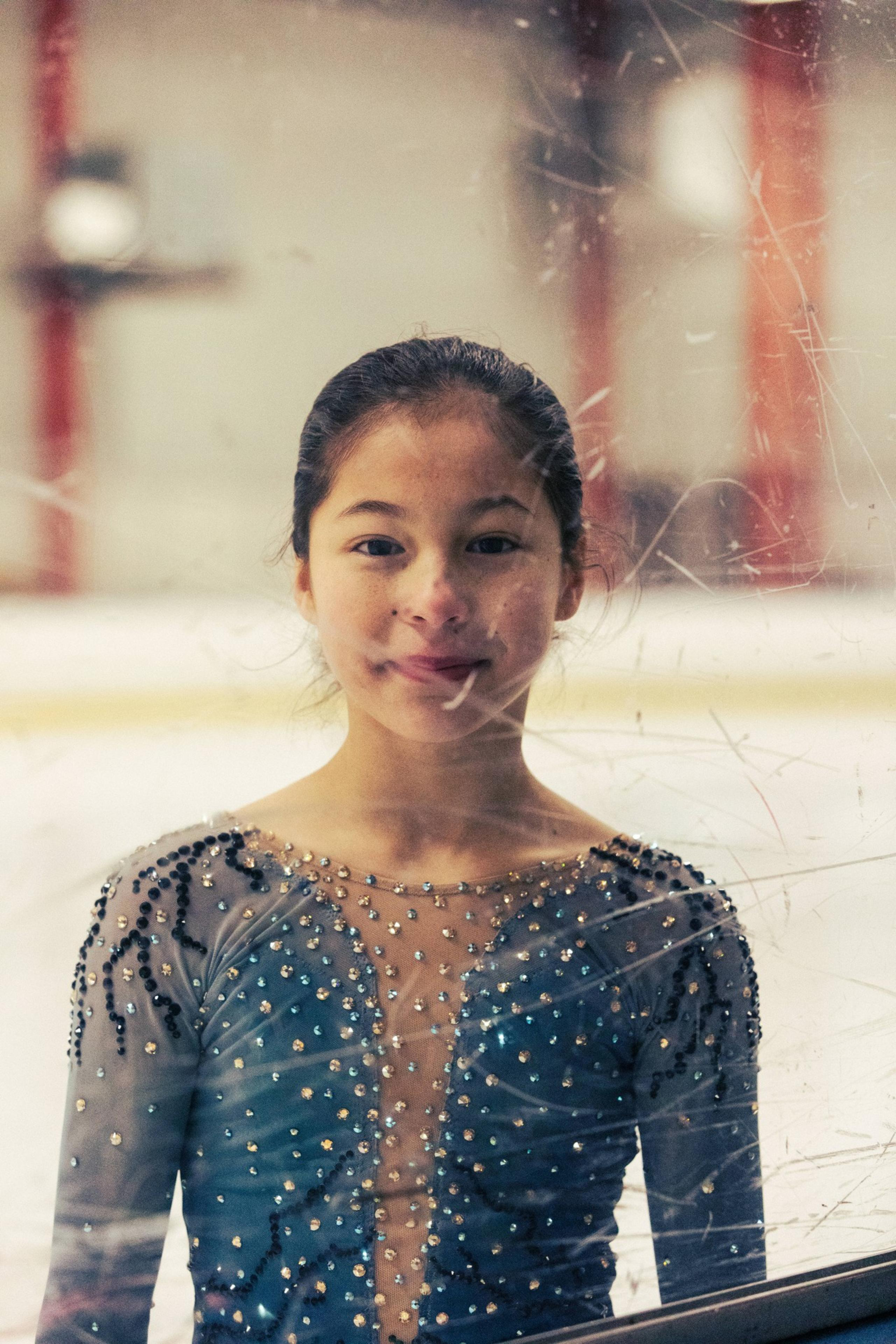 A young figure skater in a blue, rhinestone-studded costume stands behind scratched glass at an ice rink.