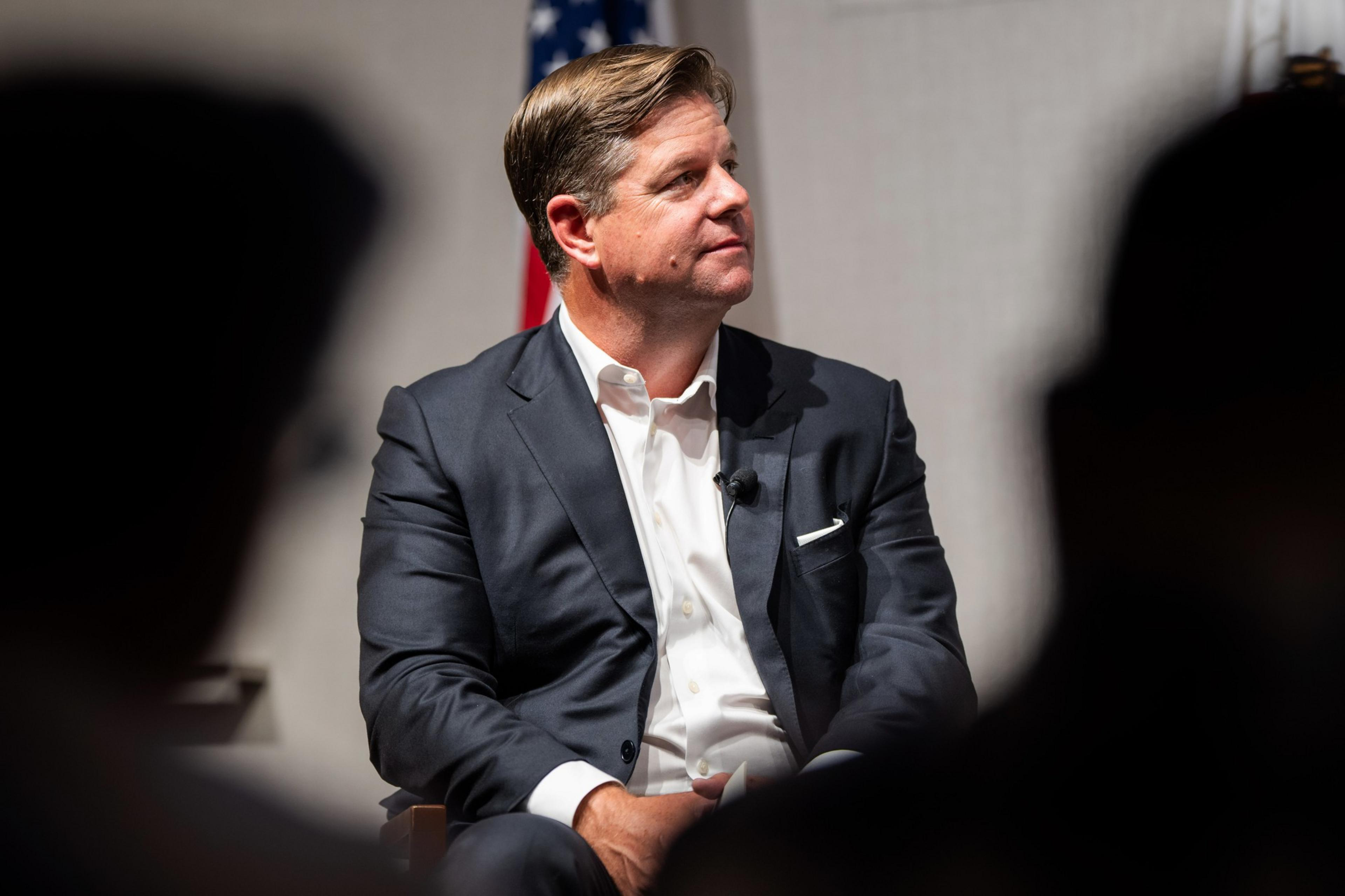 A man in a dark suit and white shirt is seated, looking to his left. He is wearing a microphone and appears attentive. A blurred U.S. flag is visible in the background.