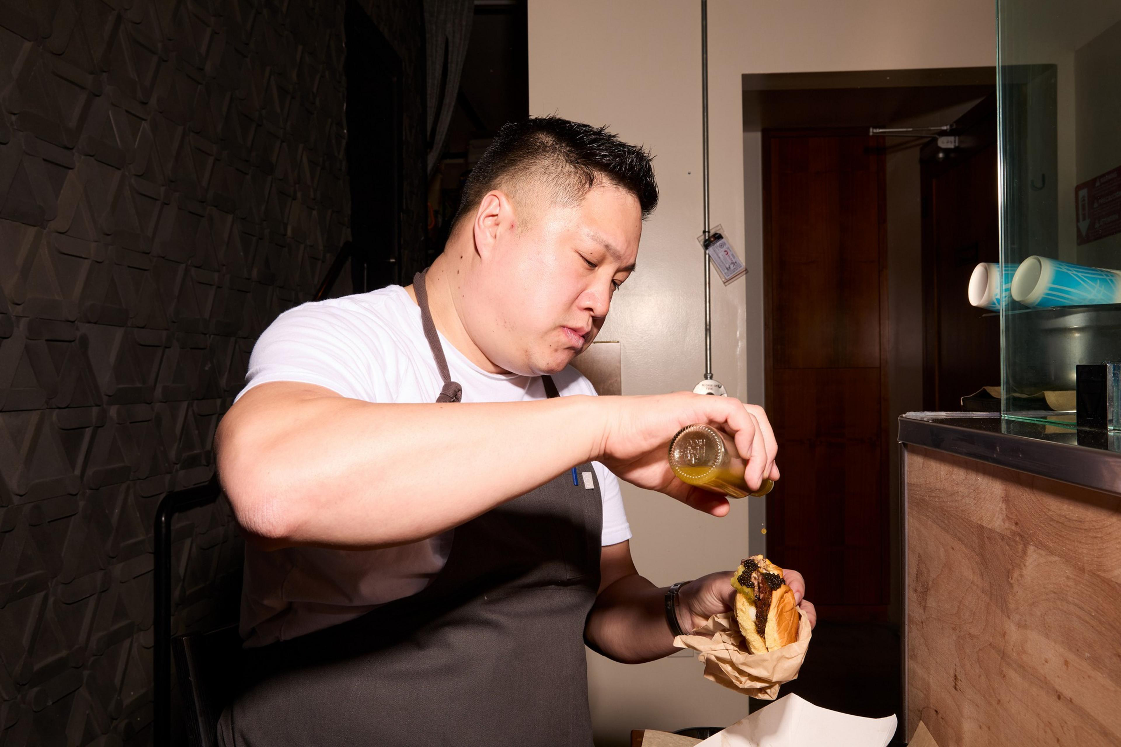 A man in an apron is concentrating as he pours a yellow sauce onto a sandwich he's holding with a paper wrap in his other hand, in a warmly lit room.