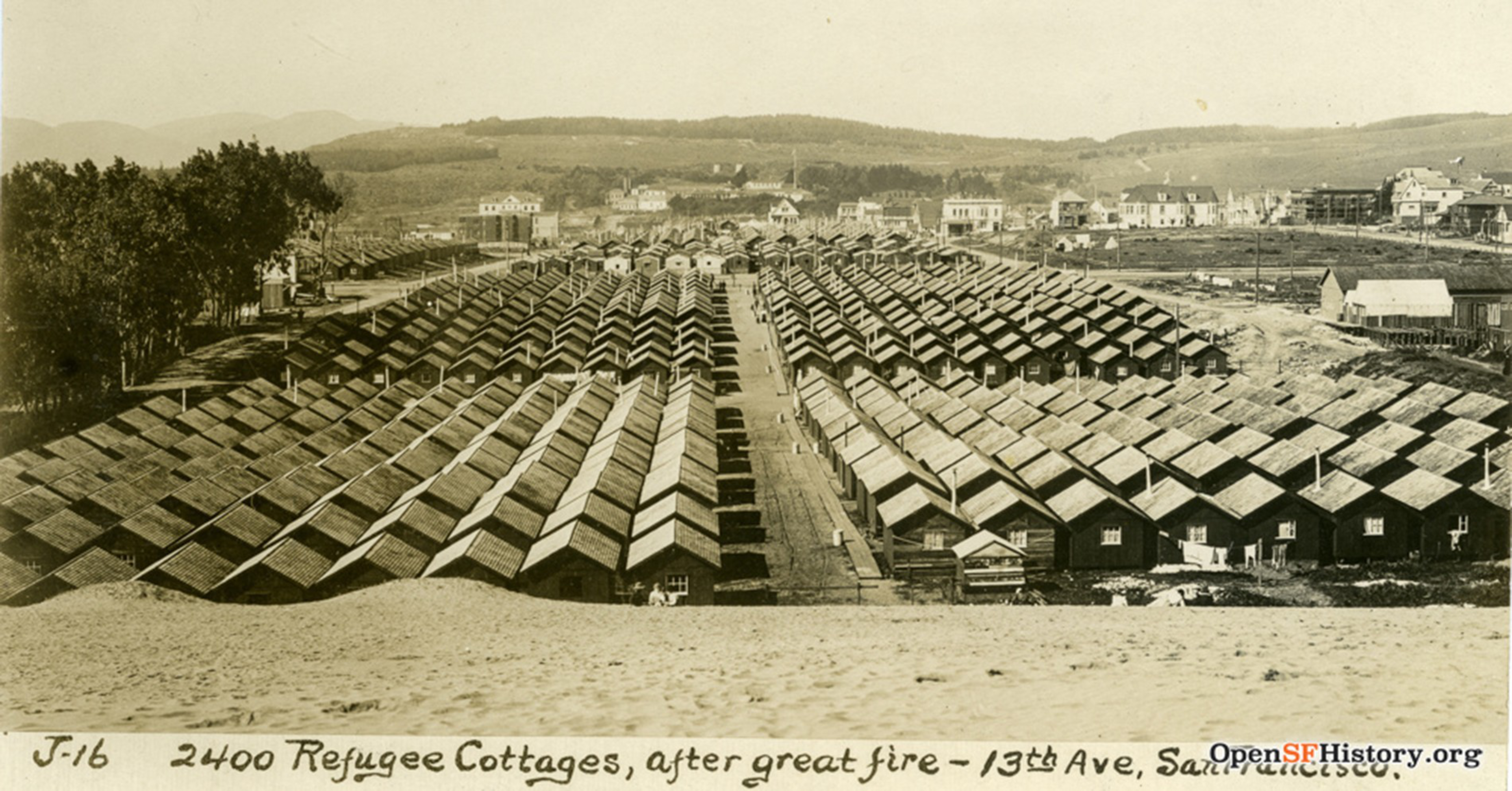 Rows of small wooden refugee cottages tightly packed, stretching back toward a town with hills in the distance, labeled after a great fire on 13th Ave.