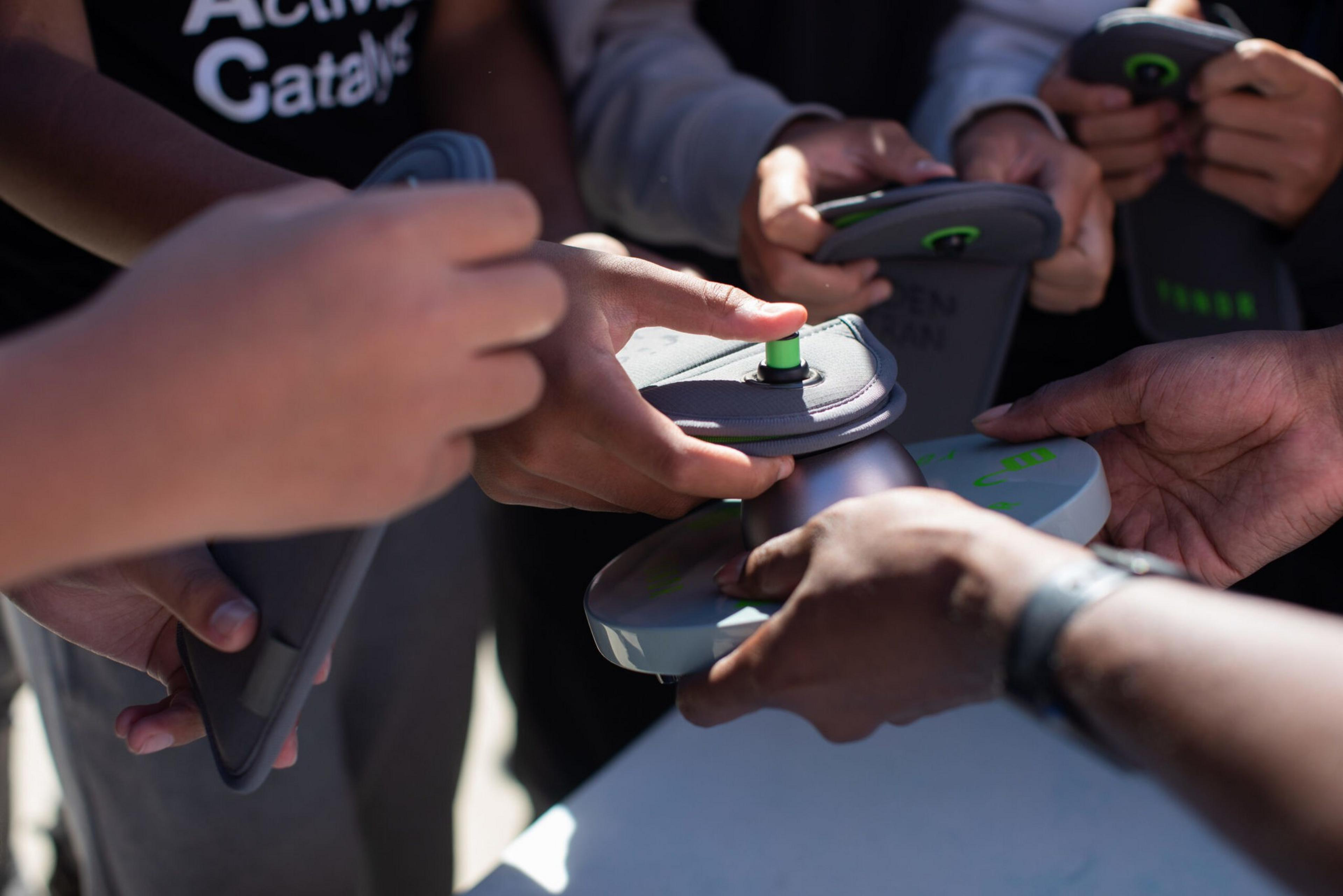 Hands are holding gray devices with green buttons, pressing them onto a circular base held by another person.