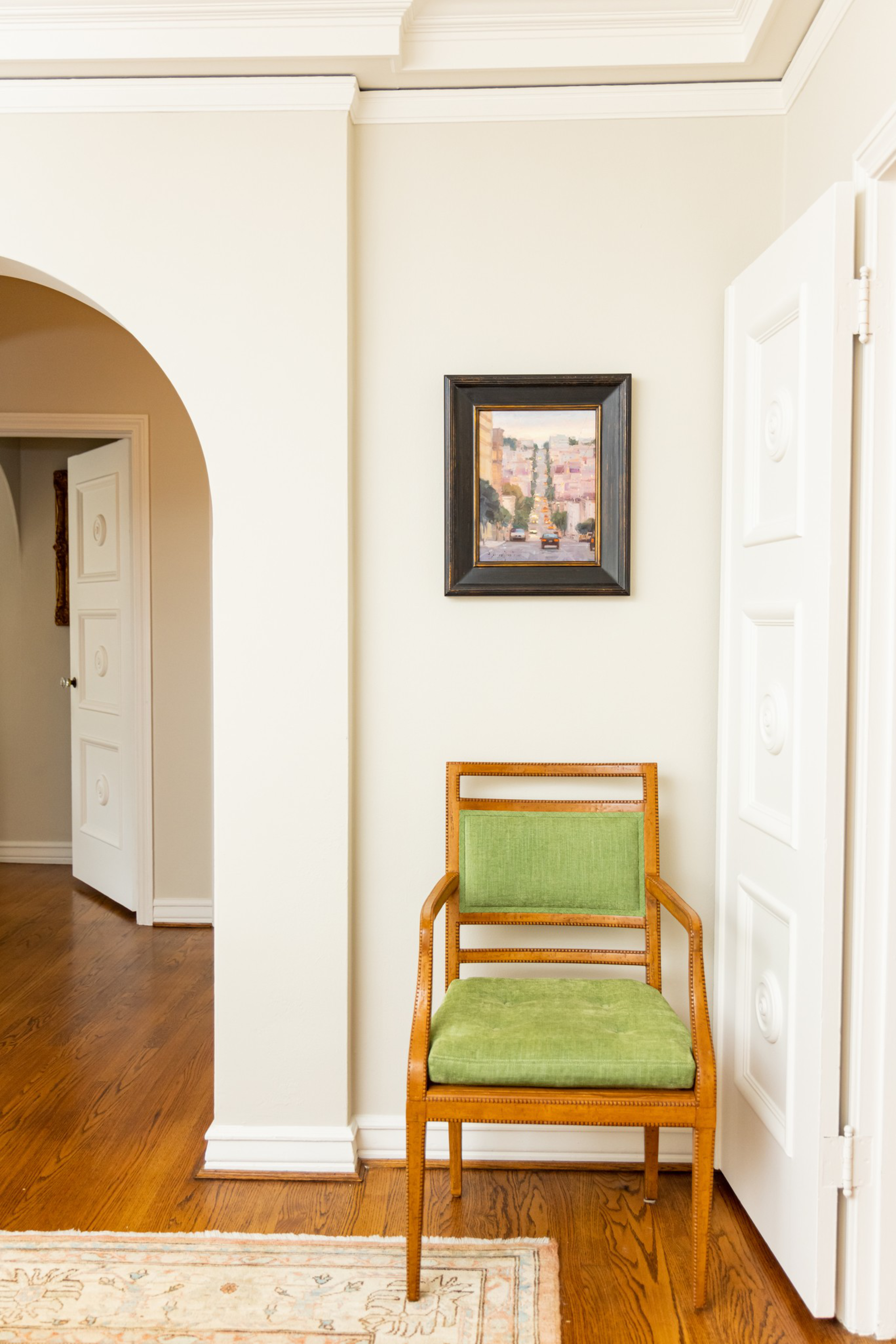 A wooden chair with green cushions sits against a white wall beneath a small framed painting, with hardwood floors and white doors nearby.
