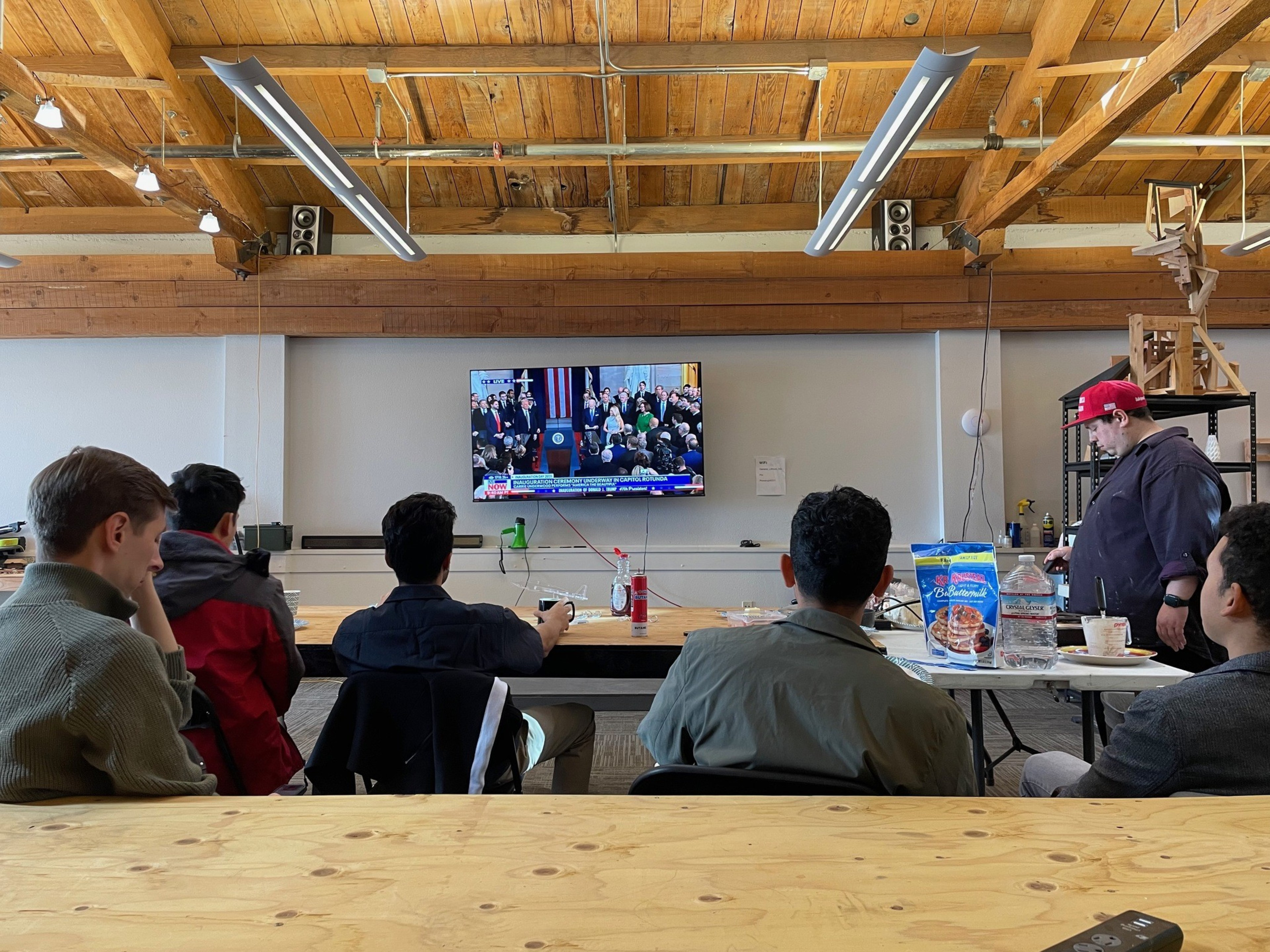 A group of people in a room with wooden beams watch a news event on a wall-mounted TV. Pancake mix and syrup are on a table with other items.