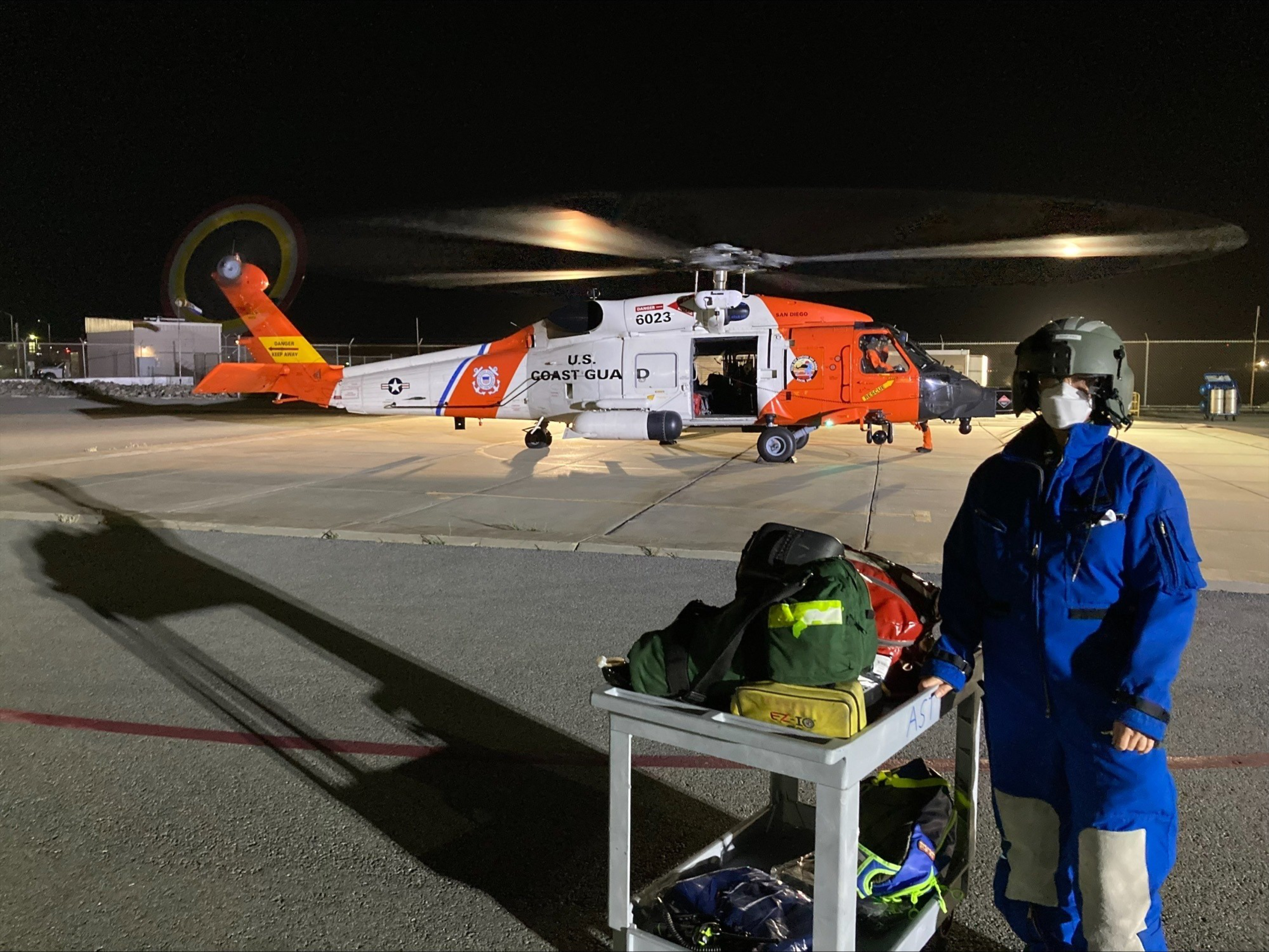 A U.S. Coast Guard helicopter with spinning rotors sits on a tarmac at night, while a person in blue flight gear and helmet stands next to a cart loaded with gear.