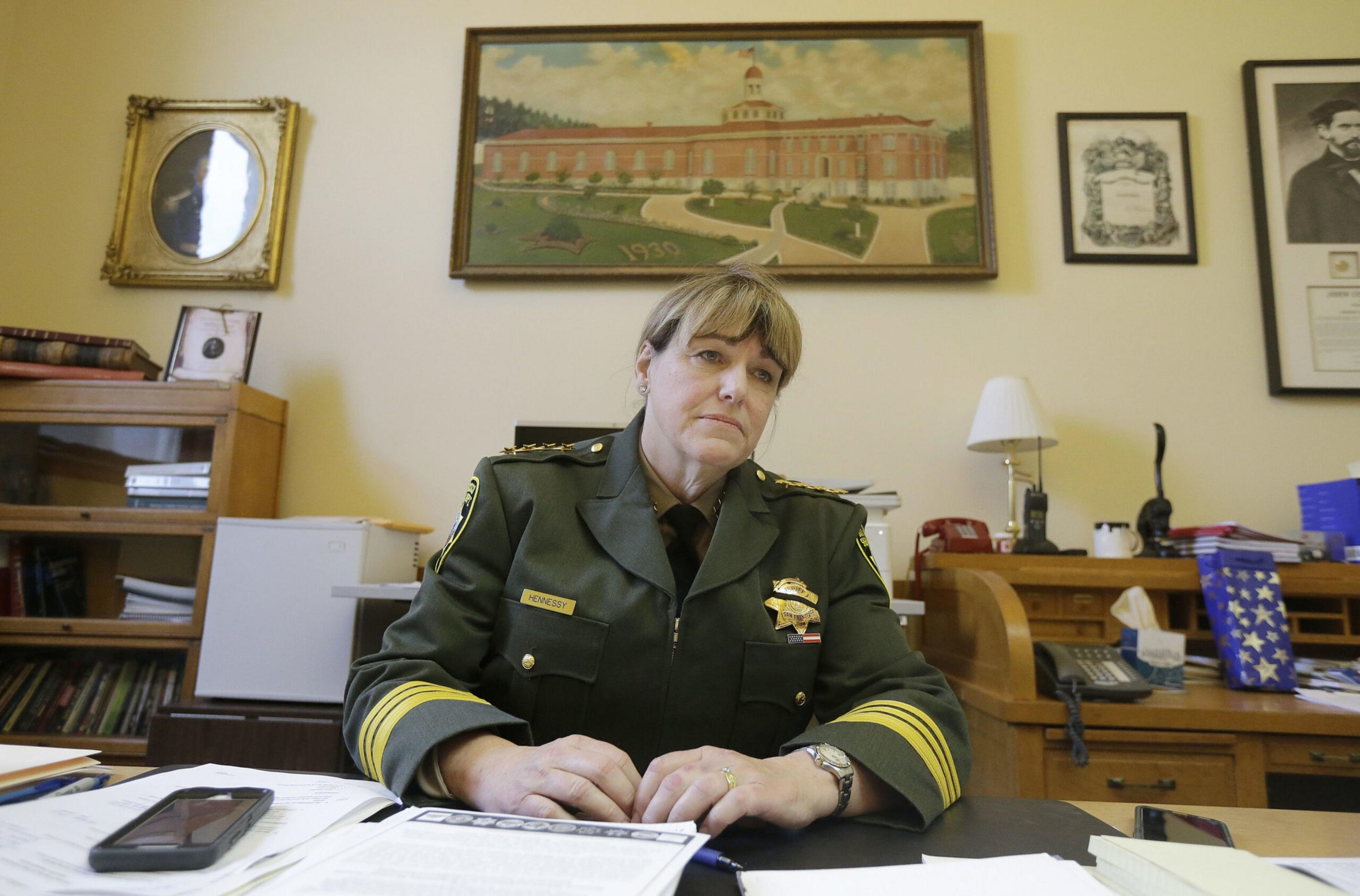 A woman in a green sheriff's uniform sits at a desk cluttered with papers and a phone, with framed pictures and a painting on the wall behind her.