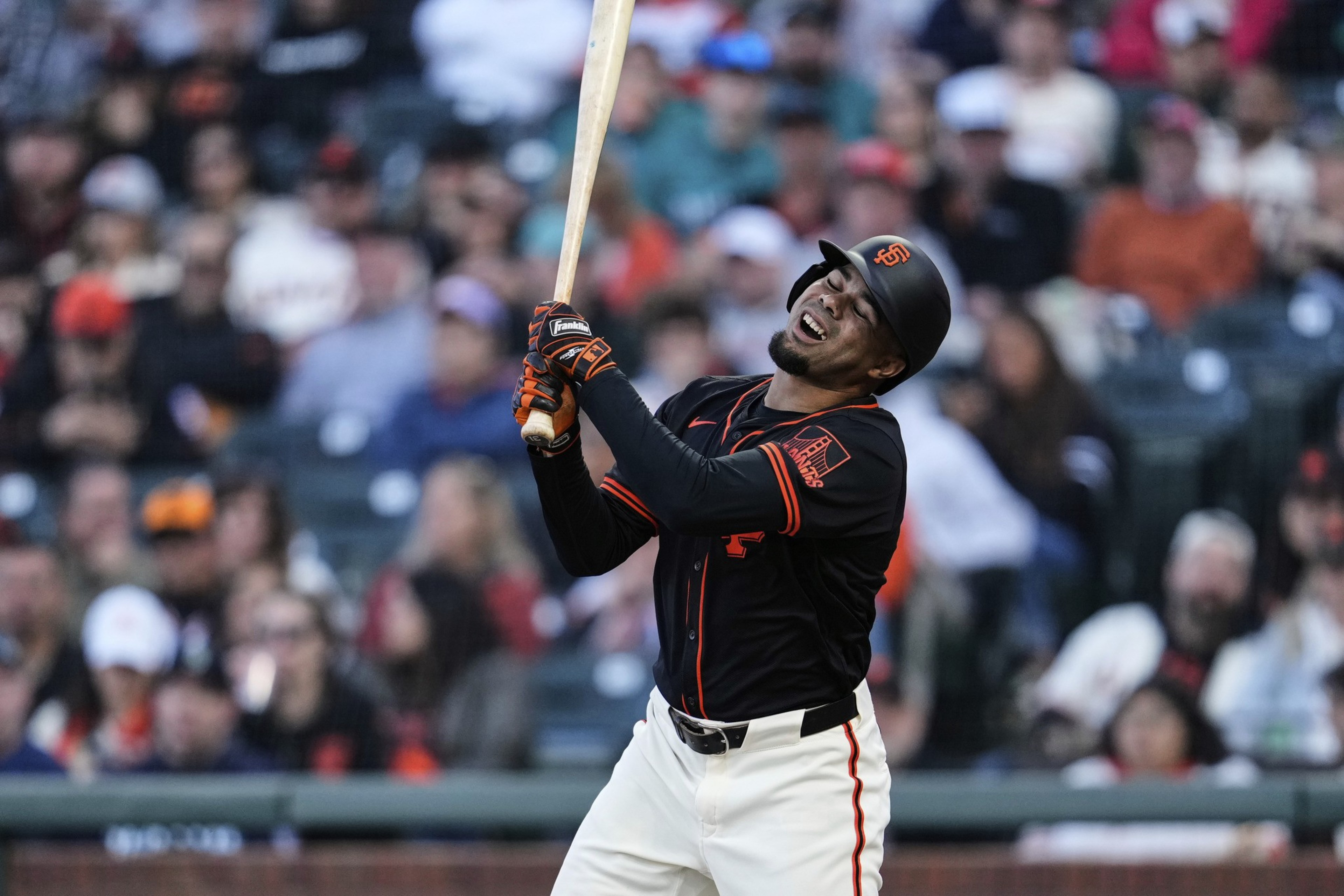 A baseball player in a black and orange uniform is mid-swing with a pained expression. He wears a helmet and gloves, and the crowd is blurred in the background.