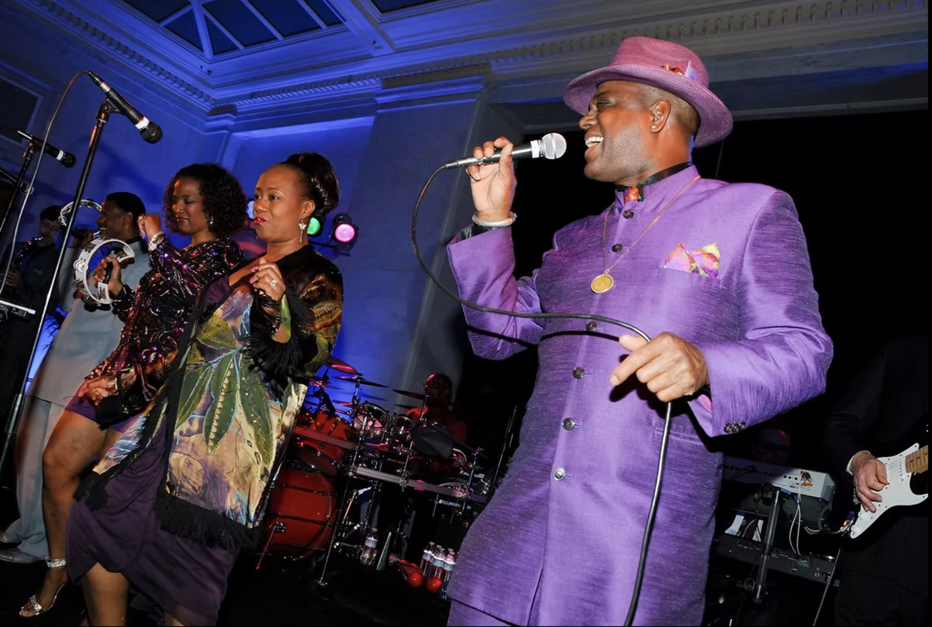 A man in a purple suit and hat passionately sings into a microphone while dancers and musicians perform alongside him on stage.