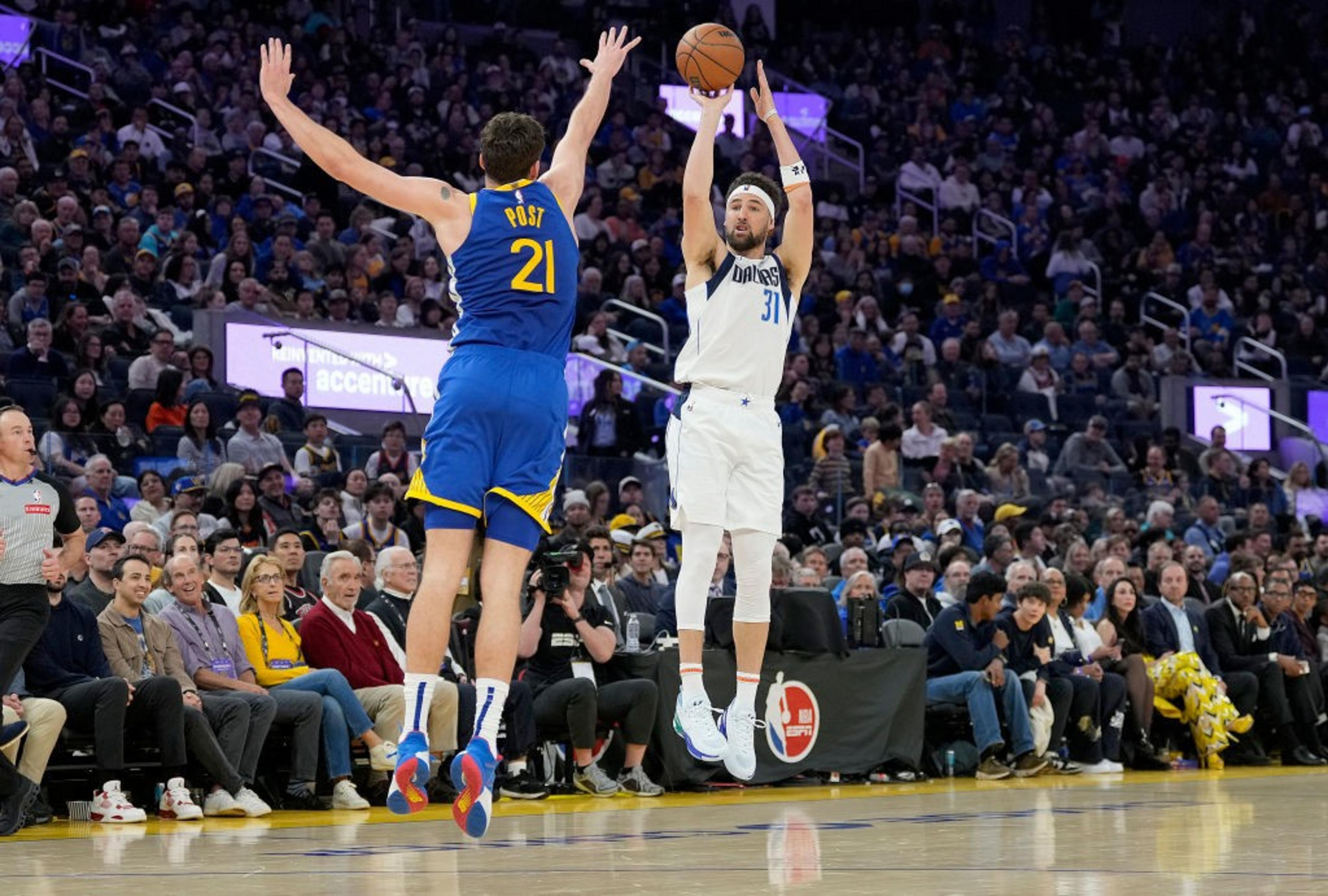 Two basketball players are in action on the court. One in white is shooting the ball, while the other in blue is jumping to block. A large crowd watches intently.