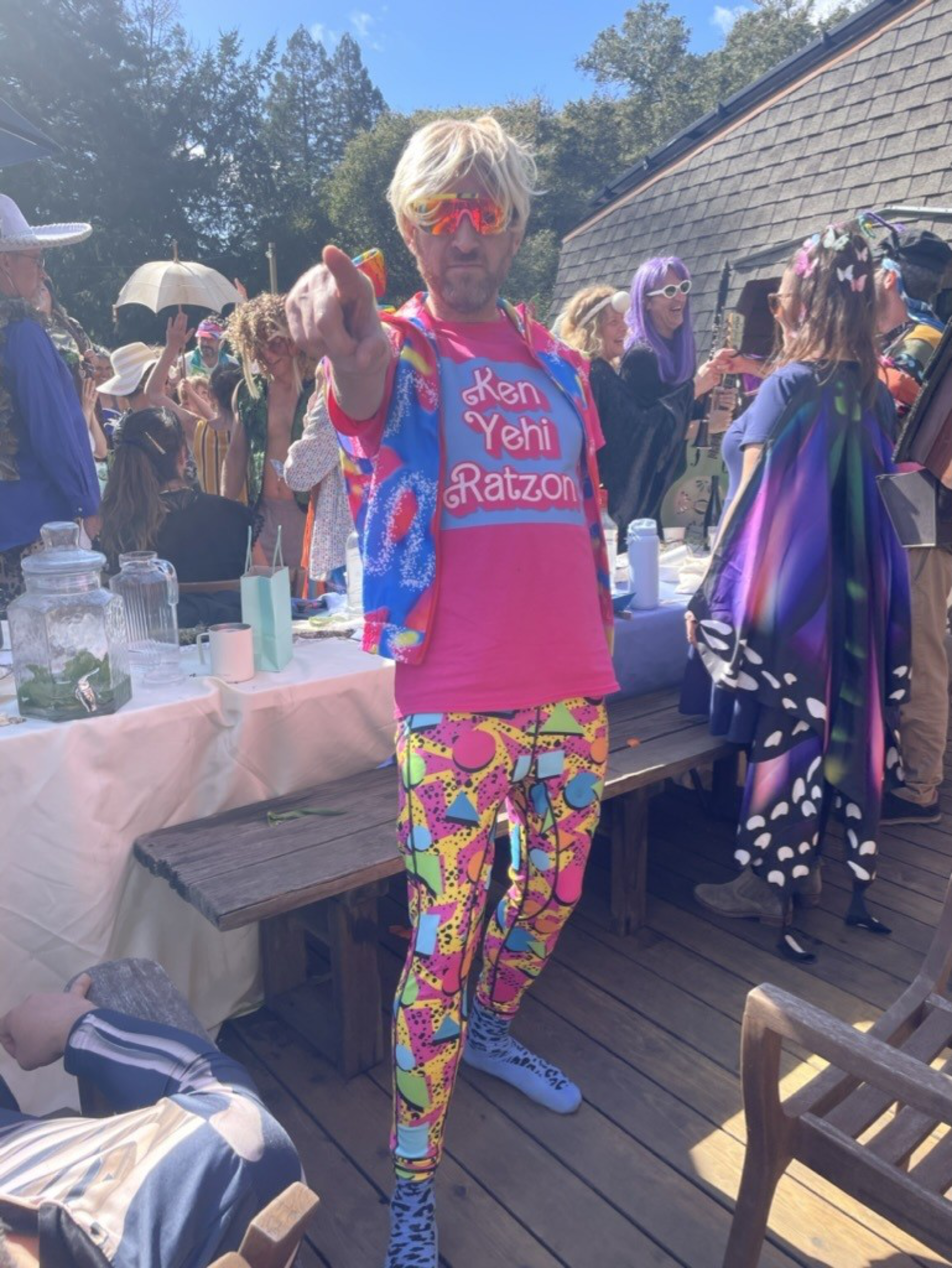 A person with blonde hair, colorful leggings, a bright shirt, and sunglasses points towards the camera at a lively outdoor gathering with similarly dressed people.