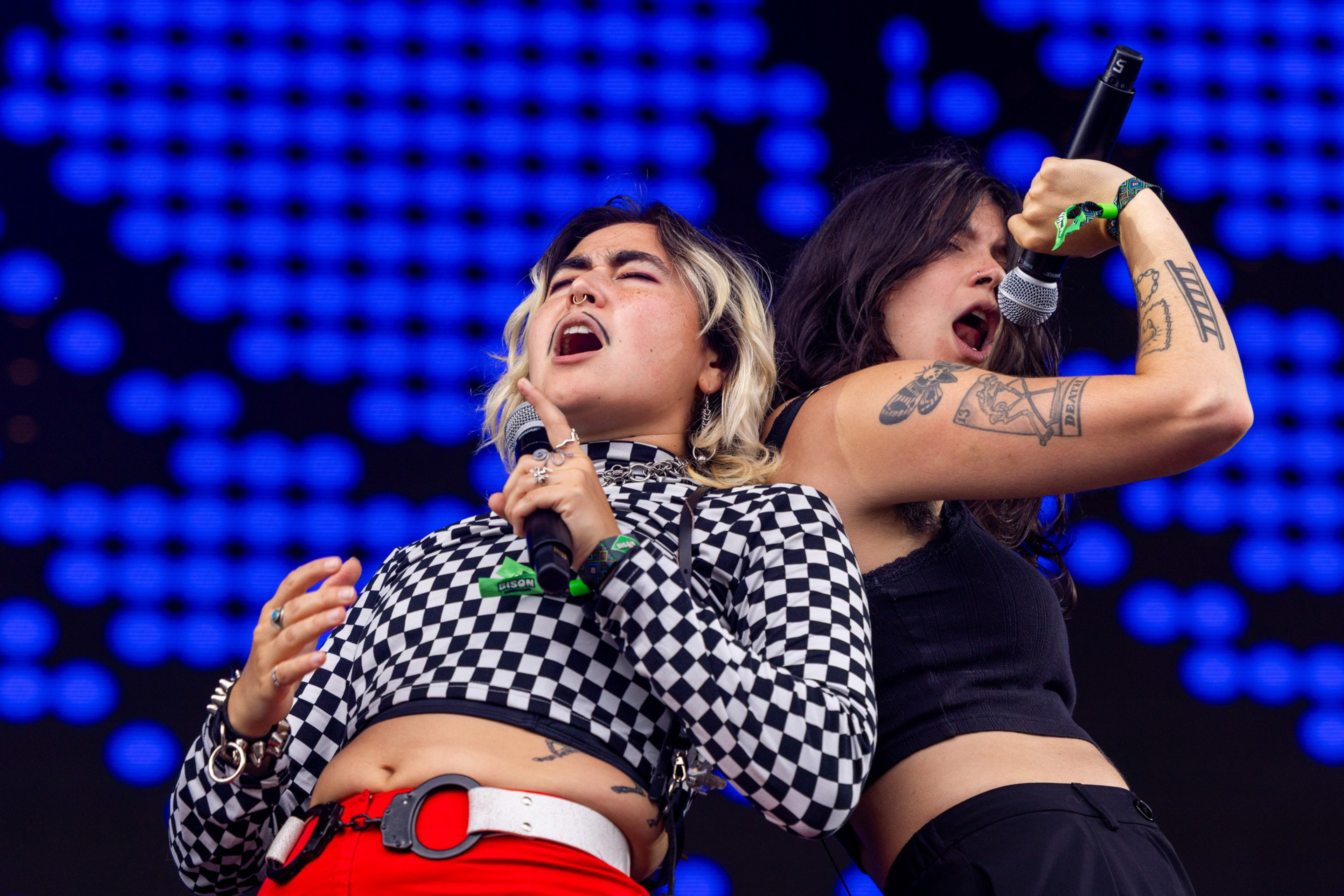 Two singers passionately perform on stage, one in a black and white checkered top and red pants, the other in a black crop top with visible tattoos.